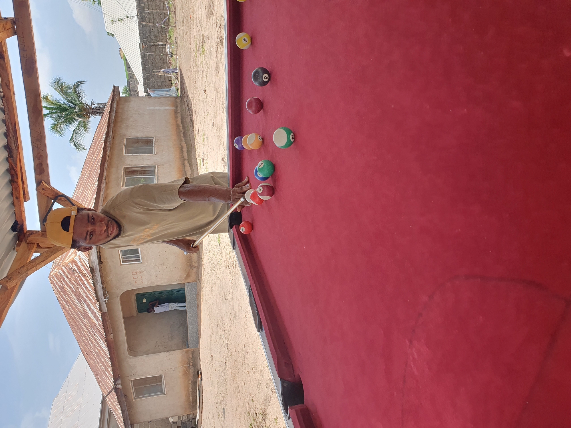 The image shows a young Black man with a yellow cap and a light-colored t-shirt playing pool. He is holding a cue stick and appears to be mid-shot. The pool table is covered in red felt and has several colored balls scattered on it, including a striped ball with the number 15 and a solid red ball with the number 3.
The scene is outdoors, with a rustic building in the background. The building has a corrugated metal roof and a door with a green frame. A palm tree is visible to the left of the building. The ground appears to be dirt or sand. The sky is blue and clear, suggesting it is daytime, likely a sunny day.
The man's expression is focused, indicating he is concentrating on the game. The overall setting suggests a casual, perhaps rural or semi-urban environment. The presence of the pool table outdoors is a notable detail. No visible text is present in the image. The image seems to be taken in Lokoja, Nigeria, given the context provided.