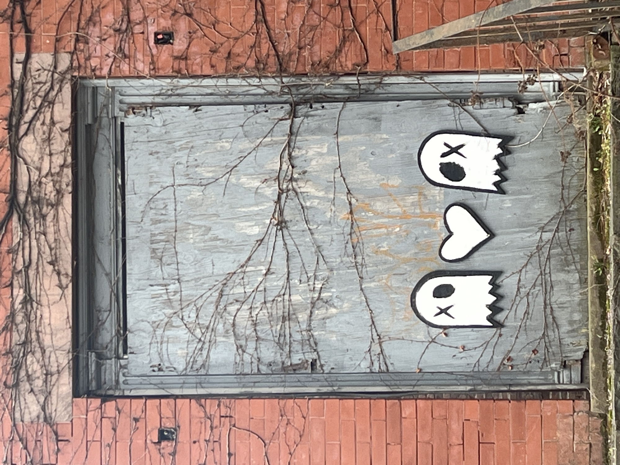 The image shows a boarded-up window in a brick building in the city of Newburgh, United States of America. The window is painted gray and features two ghost-shaped figures and a heart drawn on the surface. Vines are growing on the brick wall around the window.