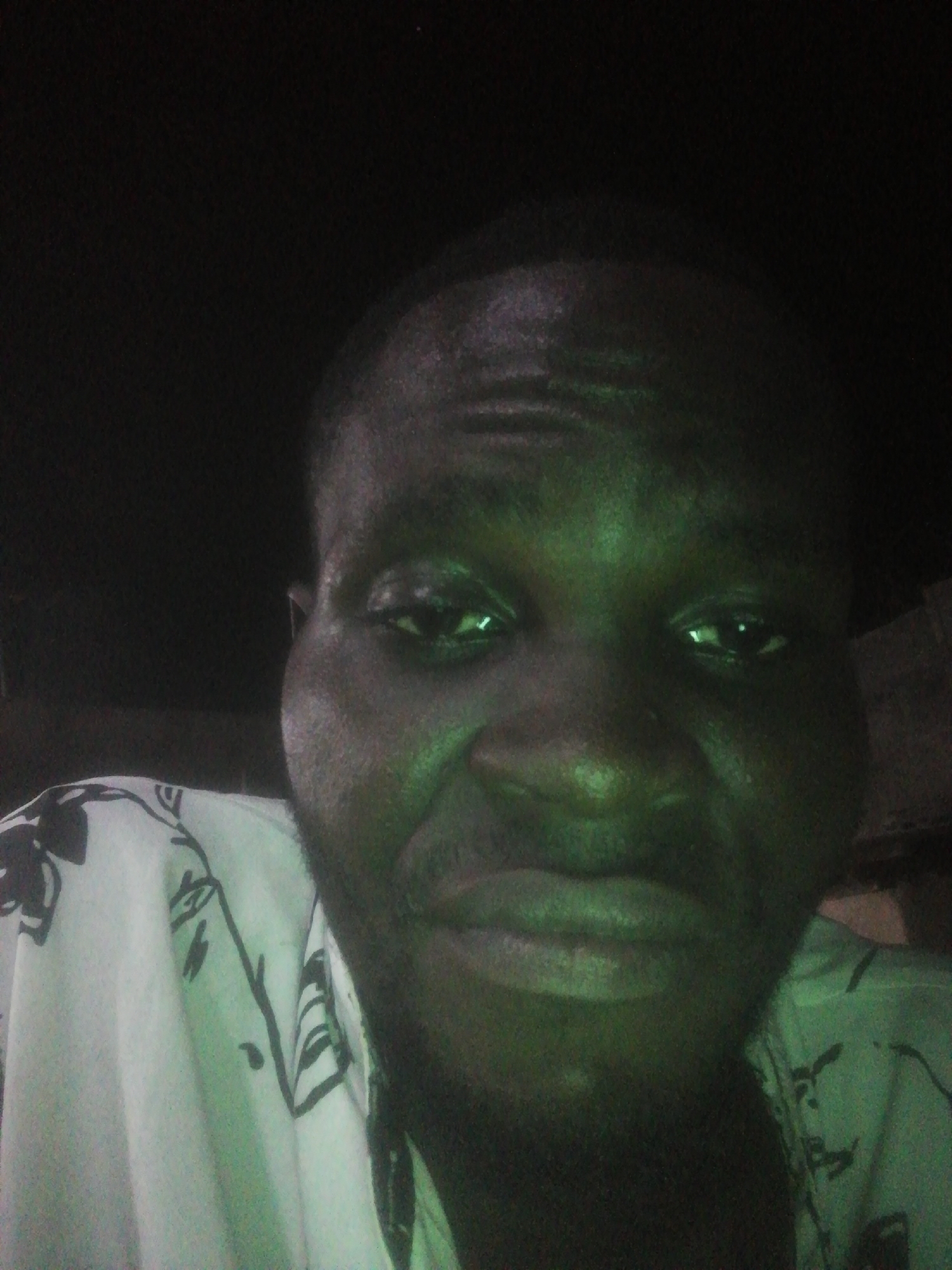 A close-up selfie shows a dark-skinned man in dim lighting. He has a shaved head and dark eyes. He is wearing a white shirt with black floral designs. The background is dark. No specific details indicate the city of Ikorodu, Nigeria.