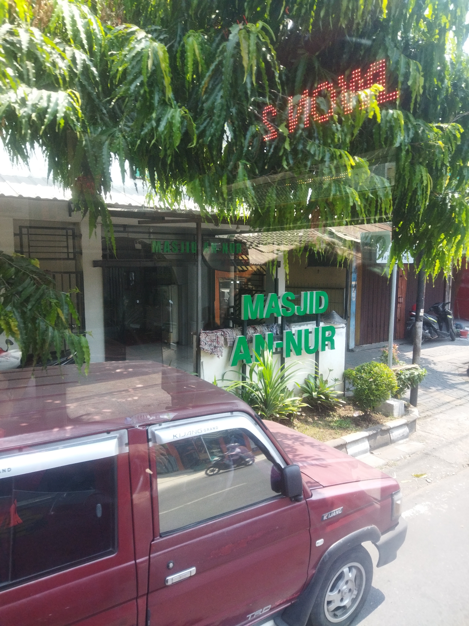 In the city of Surakarta, Indonesia, a maroon car is parked in front of a building that appears to be a mosque named "MASJID AN-NUR". The mosque has green signage and is partially obscured by the branches of a large tree with lush green foliage. A red LED sign is visible through the foliage, displaying text that reads "SMOKE". The scene is set on a sunny day, with shadows cast on the ground and the car. The car has chrome trim along the windows and a "KIJANG GRAND" decal on the passenger side. A sidewalk and a street are visible in the background, with a motorcycle parked near the mosque entrance.