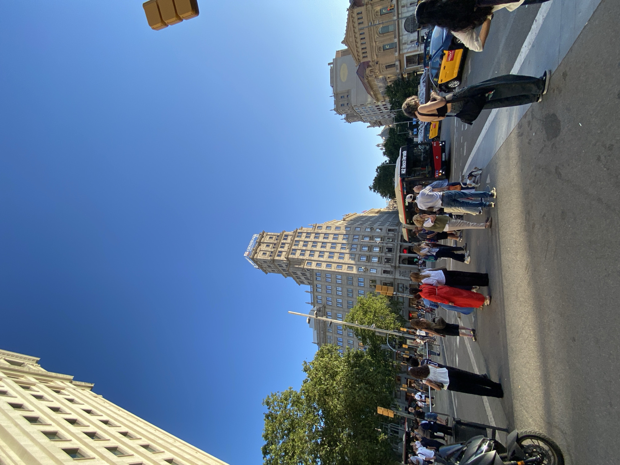 The image captures a vibrant, sunlit city street scene in Barcelona, Spain, on a clear day. The perspective is tilted, showing a wide expanse of bright blue sky dominating the upper part of the frame.
Numerous people are visible on the street and sidewalk, some appearing to wait at a crosswalk. Their attire is casual, suggesting everyday urban activity. A person in black pants and a black top stands near the center-right, next to a white crosswalk marking. Further along, a person is seen wearing a distinctive long red garment.
The street itself shows signs of urban life, with a red and white bus clearly visible, displaying the route "H12 Besòs Verneda." A yellow and black taxi is also parked or moving in the background, a common sight in Barcelona. A black motorcycle is partially visible in the lower right foreground. Traffic lights, primarily showing yellow, are mounted on poles.
Architecturally, the scene features several multi-story buildings. A particularly tall, ornate building with many windows dominates the left side of the frame when oriented correctly. It has "GENERALI" signage prominently displayed near its top, indicating an insurance company. Other buildings of varying heights and styles line the street. Trees with green foliage add a natural element to the urban landscape.
The bright sunlight and clear sky indicate it is daytime, likely mid-morning or afternoon, with pleasant weather conditions. The overall impression is that of a bustling European city, with people and traffic moving through its historic streets.