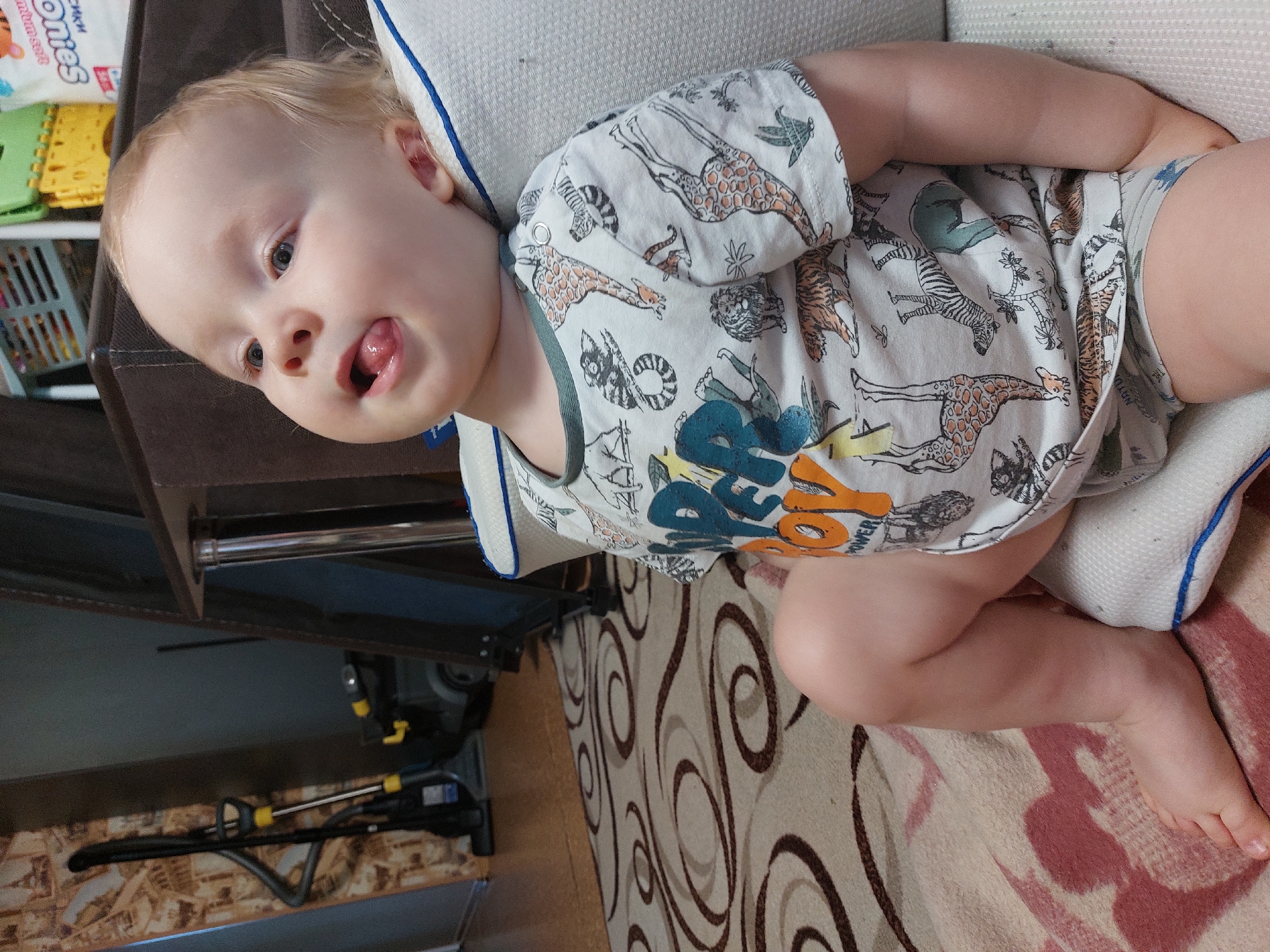 A baby wearing a light-colored onesie with animal prints lies on a light-colored cushion with blue trim on a patterned rug. The baby is sticking out its tongue. A brown sofa is partially visible in the background. A vacuum cleaner is visible on the floor, alongside wooden flooring. The image was taken indoors in Chaikovsky, Russia.