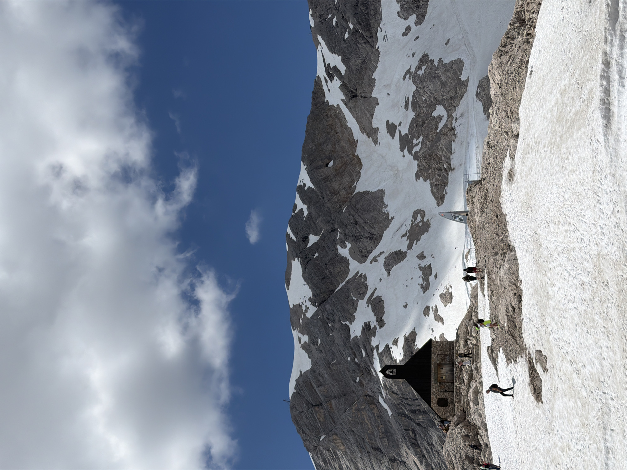 The image depicts the peak of the Zugspitze, Germany's highest mountain, located near Garmisch-Partenkirchen. A small, dark-roofed building, resembling a chapel or information center, is situated on a rocky outcrop covered in snow. Several individuals are present around the building and on the snowy slopes, some appearing to be tourists. The mountain behind is rugged and partially covered in snow, with a clear blue sky above featuring scattered white clouds. The scene suggests a high-altitude, mountainous environment with remnants of snow despite the sunny weather.