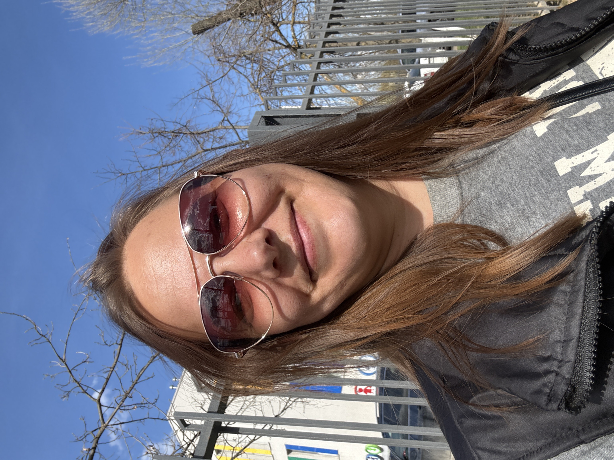 A woman with brown hair, wearing sunglasses, a grey shirt, and a black jacket, is posing for a selfie outdoors. The background includes a grey metal fence, bare trees, a clear blue sky, and a building with visible signs, possibly indicating a station or service area in the city of Dmitrov, Russia.