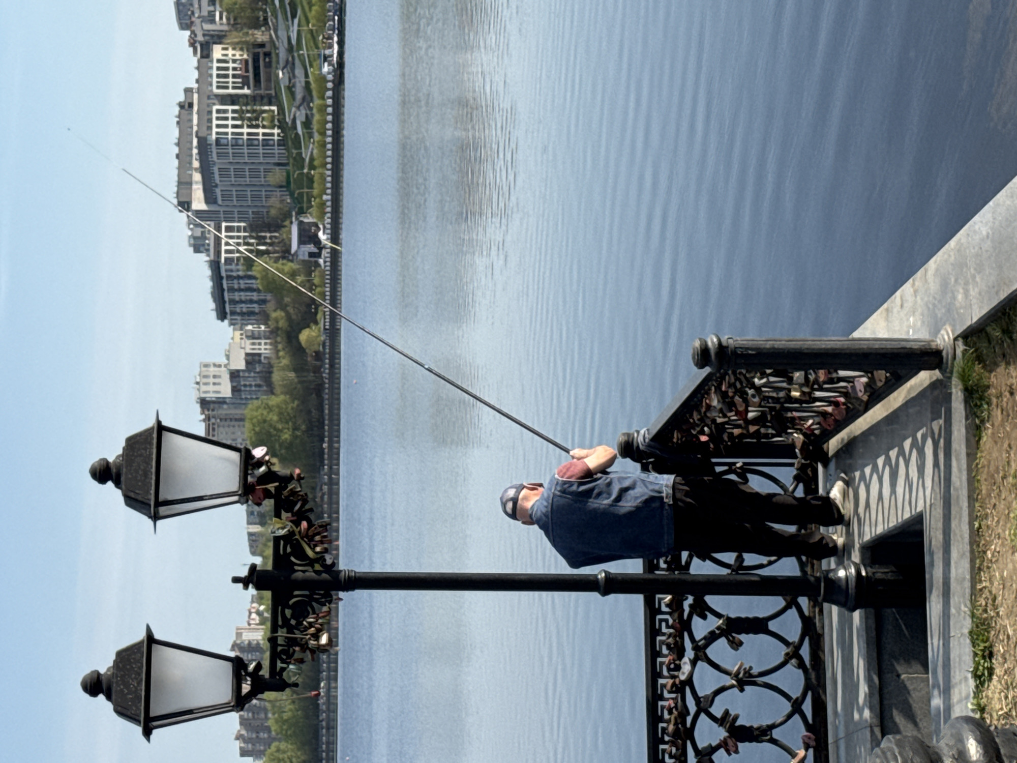 An individual is fishing from a quay in Izhevsk, Russia. They stand near a black ornate fence, which is adorned with numerous padlocks. Adjacent to the fence is a tall, black lamppost with two lamps. The fishing line extends out over a body of water that spans most of the frame. Across the water, buildings and greenery can be seen. The sky is blue and appears clear.