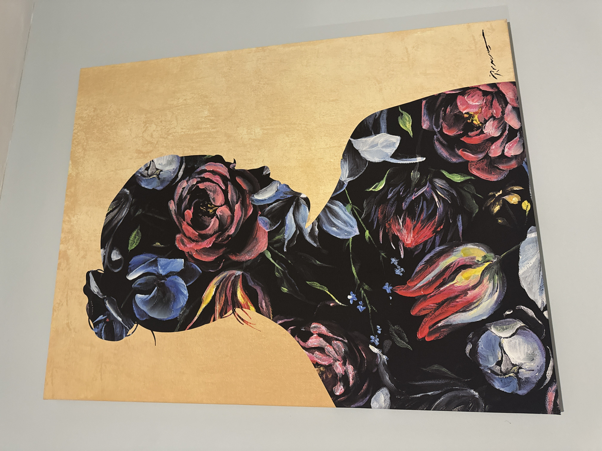 The image shows a painting hanging on a light grey wall. The painting features a gold background with the silhouette of a woman's head and shoulders filled with floral patterns in dark colors like black, red, and blue. The floral patterns include roses and tulips. There is a signature in the upper right corner of the painting. The location is unknown.