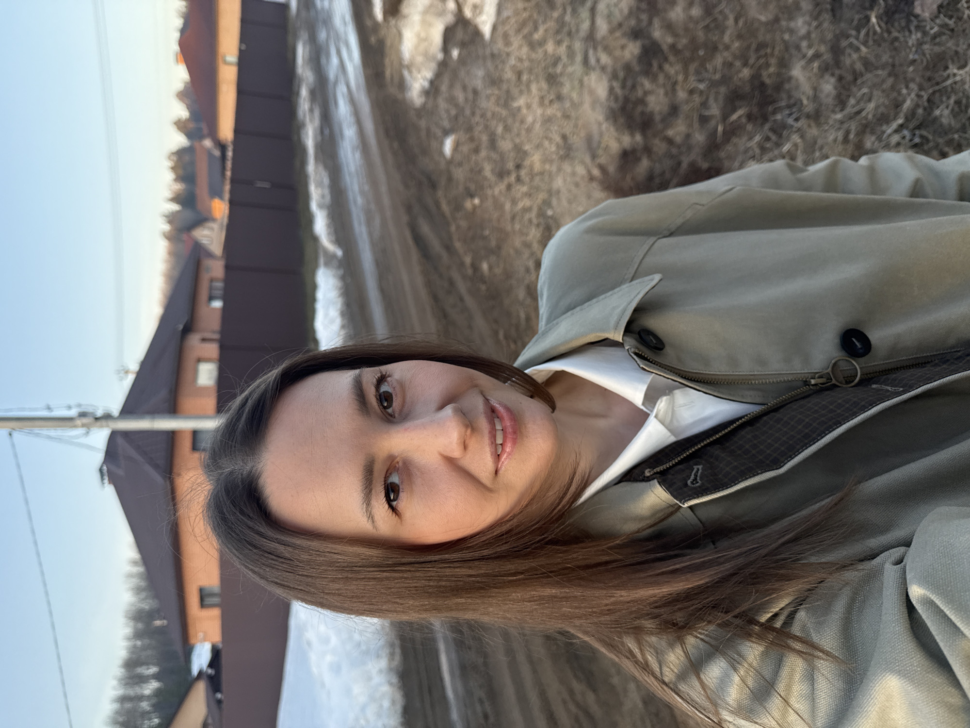 A woman poses for a selfie outdoors. She has long brown hair and is wearing a white shirt under a light green jacket. The background includes residential buildings and a snowy or slushy road. The location appears to be Chaikovsky, Russia, based on visual cues like the architectural style of the houses and the presence of snow.