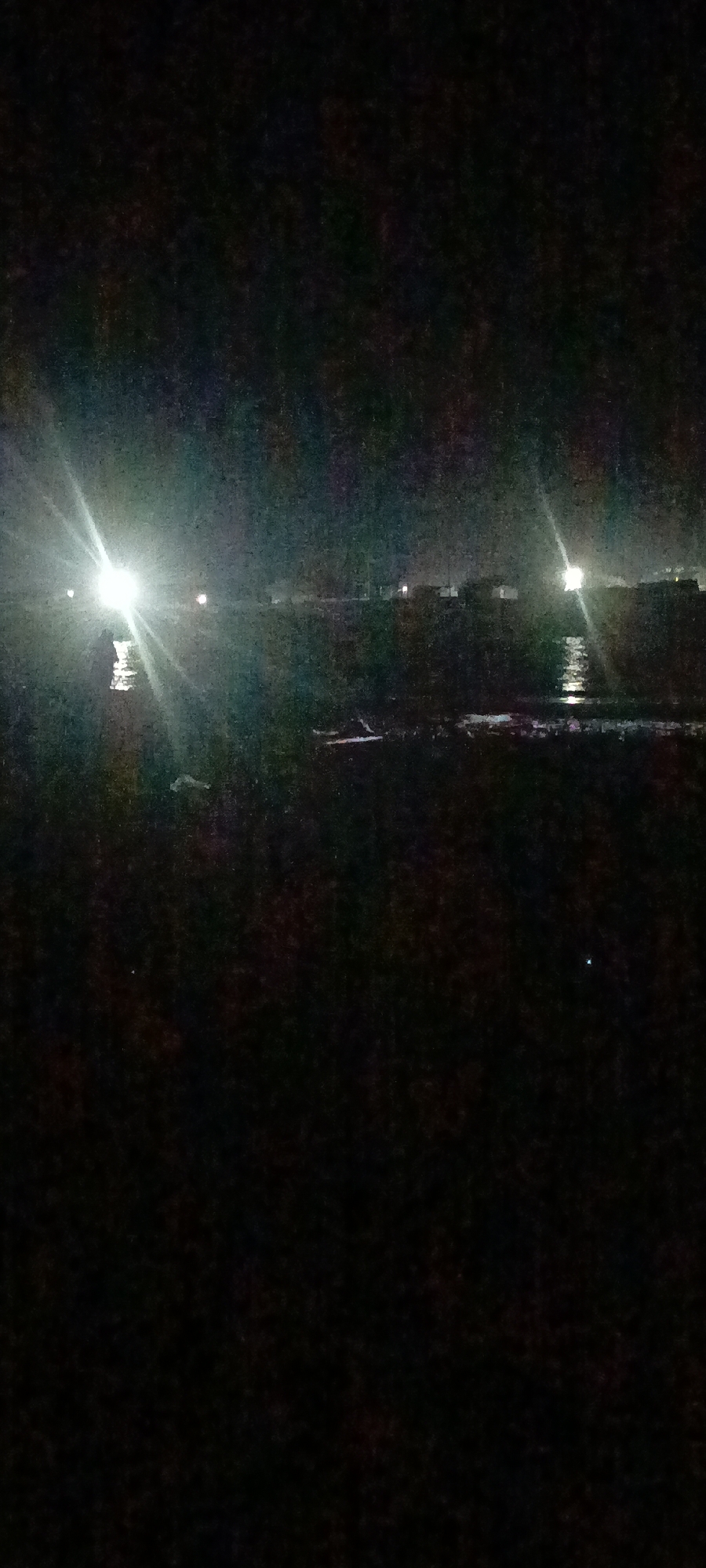 A nighttime photograph captures a dark, possibly rural or coastal scene with several bright lights. In the foreground, the ground appears dark and indistinct, with some textured patches suggesting vegetation or uneven terrain. The middle ground features a body of water, reflecting some of the light sources. Across the water, a distant line of structures or landscape is visible, punctuated by multiple bright lights. The most prominent light is a harsh, white, starburst-like illumination on the left side of the image, possibly from a vehicle's headlights or a powerful spotlight. Other lights are scattered along the horizon, some appearing as smaller, less intense glows, and one with a distinct reflection on the water. The sky is dark and largely obscured by the bright lights. The overall impression is one of a dimly lit, possibly undeveloped area at night, with artificial lighting creating points of focus.