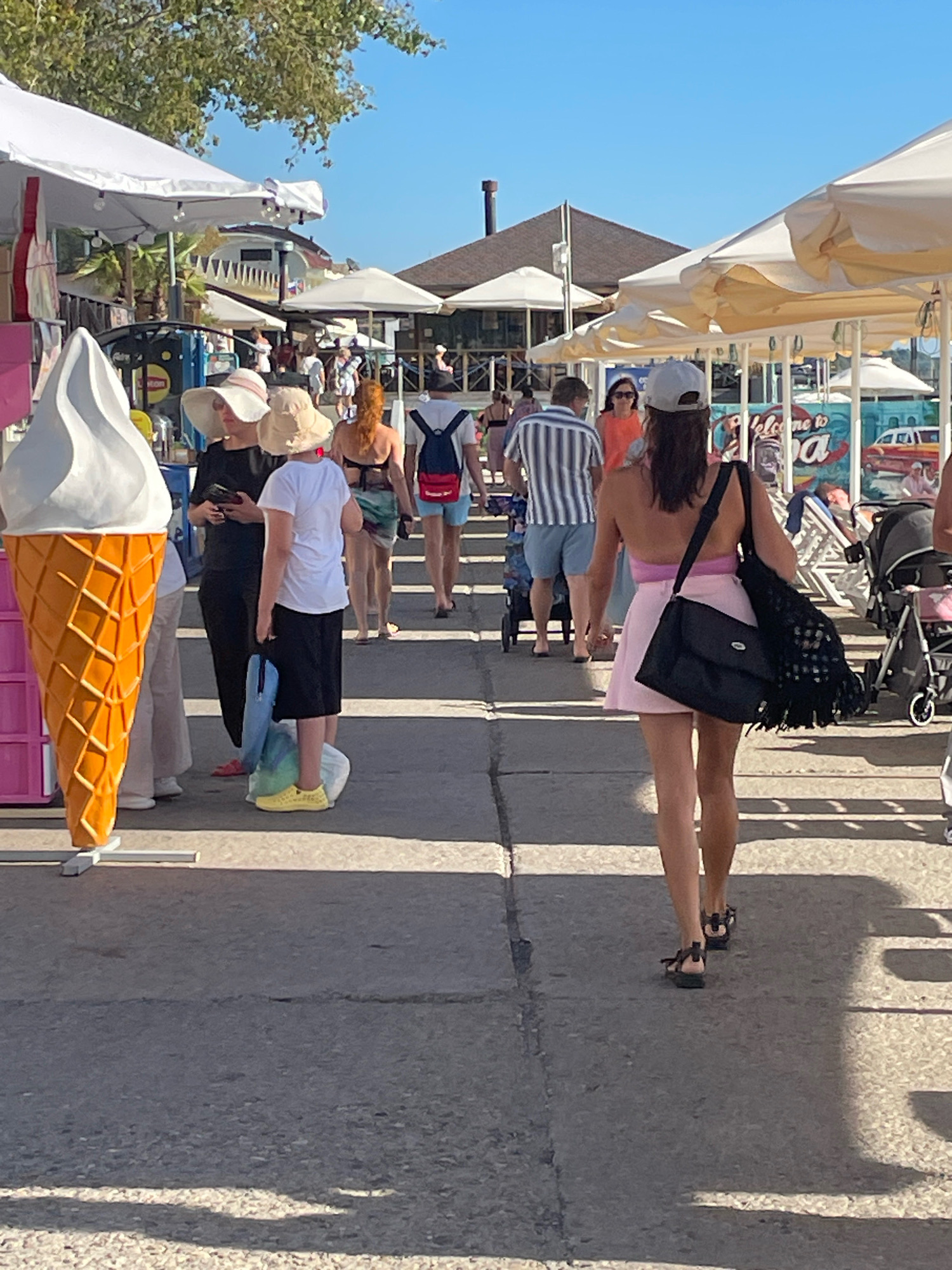 The image depicts a sunny day on a paved walkway, likely a promenade or boardwalk, in Sochi, Russia. The walkway is lined with white and cream-colored umbrellas, suggesting a commercial area with shops or cafes. A large, decorative ice cream cone sculpture stands prominently on the left.
Several people are walking along the path. In the foreground, a woman with a grey baseball cap and a pink skirt walks away from the camera, carrying a black shoulder bag. To her left, a woman in a black dress and hat holds a phone, and a person in a white shirt and black skirt stands with yellow footwear, carrying bags. Further down the path, a man in a striped shirt and blue shorts walks with a backpack, and other individuals are visible in the background, including a woman in an orange top and a woman with red hair in a swimsuit.
Various strollers and beach chairs are visible near the umbrellas, indicating a location frequented by tourists. A sign with the text "Welcome to Sochi" is partially visible in the background, confirming the location. The overall scene is one of casual activity and commerce in a coastal resort town.