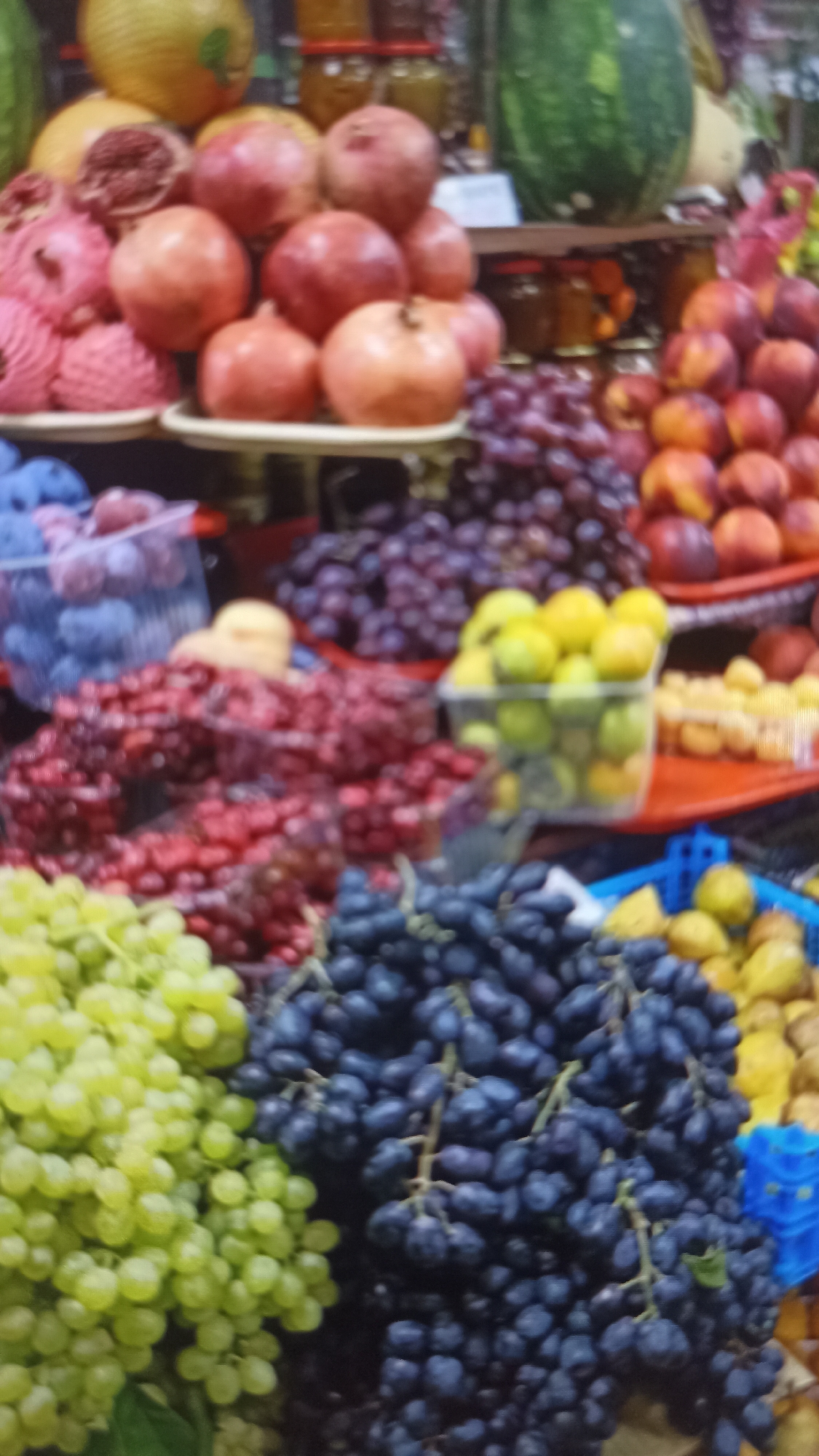 This image captures a vibrant display of fruits at an outdoor market. In the foreground, large bunches of green and dark purple grapes are prominently featured, with the purple grapes filling a significant portion of the frame. To the left, a pile of red berries, possibly cranberries or cherries, is visible in plastic containers. Behind these, there are more grapes, this time in shades of deep purple and a lighter, reddish-purple hue. Further back, a stack of bright red pomegranates and some pink-wrapped fruits, possibly also pomegranates or apples, are arranged on trays. In the background, a large green watermelon and jars of preserves or jams are stacked on shelves, suggesting a diverse selection of produce. The overall impression is one of abundance and freshness, typical of a bustling market setting. The specific location is unknown, but the scene is indicative of a local market where fresh produce is sold.