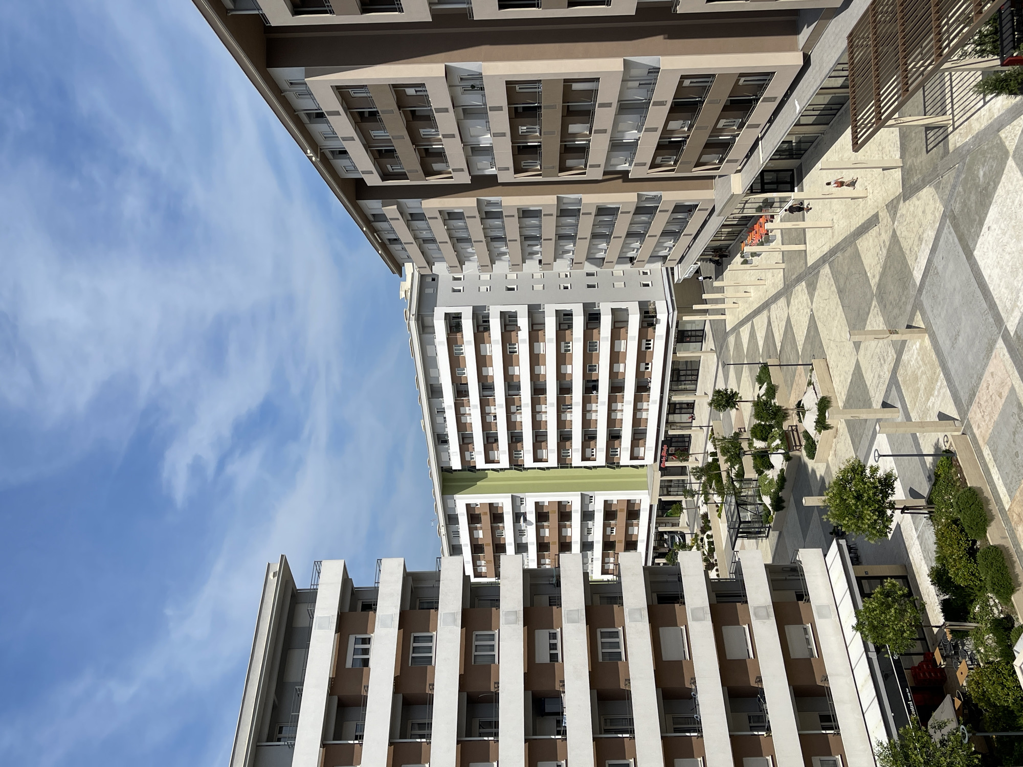 This is a high-angle, eye-level photograph of a modern residential and commercial complex in Podgorica, Montenegro, under a bright, partly cloudy blue sky. In the foreground, to the left, is a multi-story residential building with balconies featuring brown panels and white railings. Adjacent to this building on the ground floor are shops with awnings, including one labeled "Central Lounge Bar."
Further into the scene, other similar residential buildings of varying heights and architectural styles are visible. One building in the mid-ground is distinguished by a vertical, light green accent strip. The ground level of these buildings features commercial spaces with large glass windows.
The outdoor space between the buildings is a pedestrian plaza paved with geometric patterns in light and dark shades of beige and gray. Scattered throughout the plaza are small trees, manicured bushes, benches, and streetlights. A modern, small glass-enclosed structure with greenery inside is also present.
A few individuals are scattered throughout the plaza, walking or appearing to be patrons of the ground-floor businesses. The lighting suggests it is daytime, likely late morning or early afternoon, with distinct shadows cast by the buildings and structures. The overall scene depicts a contemporary urban development focused on mixed-use living and retail.