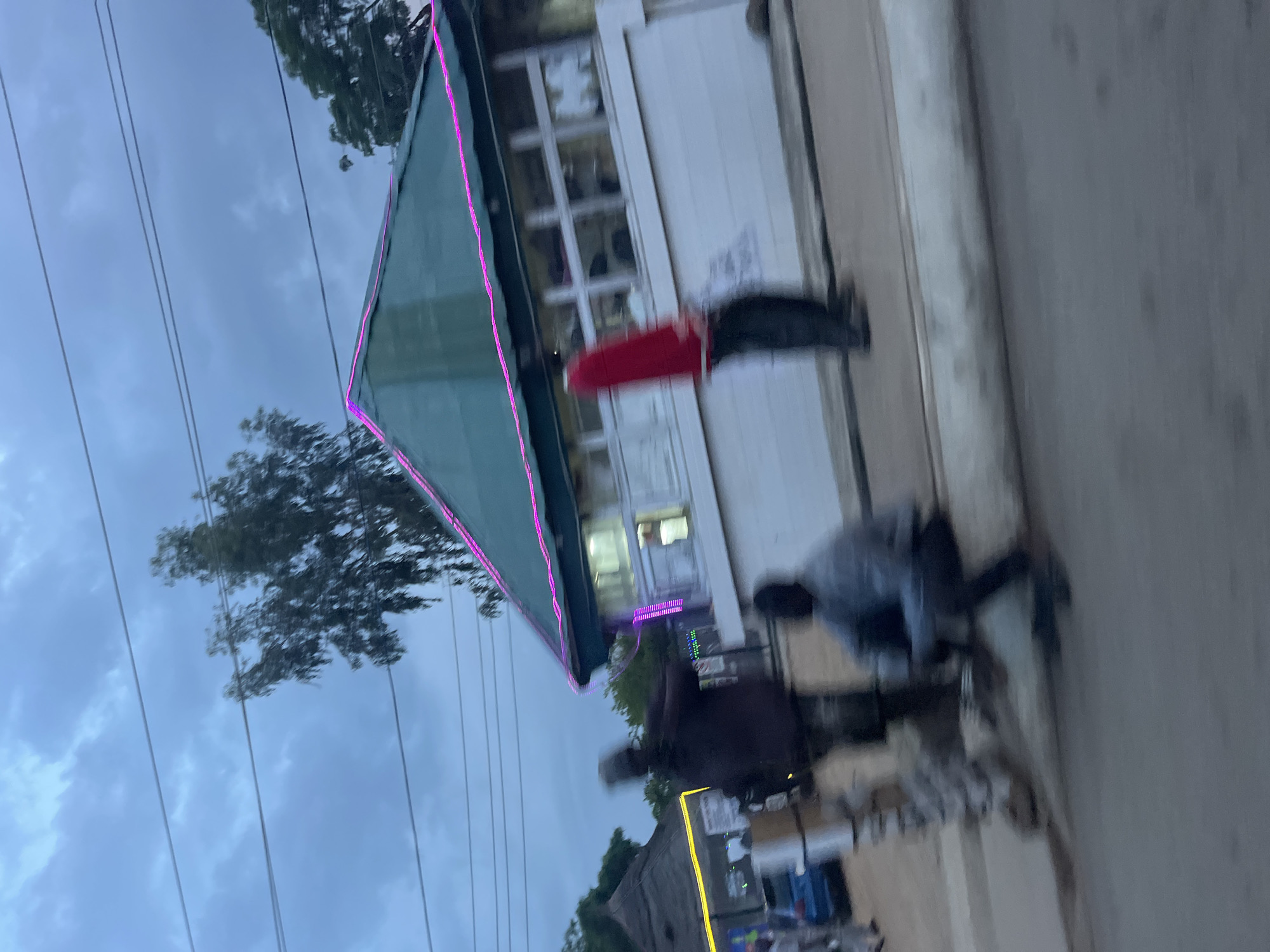 The image depicts a blurry outdoor scene, likely in Bukuru, Nigeria, during twilight. A building with a green roof adorned with pink neon lights is visible, with several windows along its facade. In front of the building, three figures are present. One person, wearing a red top and dark pants, stands upright. Another, in darker clothing, appears to be moving. A third person is seated. To the left of the main building, another structure with a thatched roof and yellow neon lights is partially visible, with a blue car parked nearby. The overall impression is one of a lively, possibly commercial, area with ambient lighting and people engaged in various activities. The sky above is overcast.