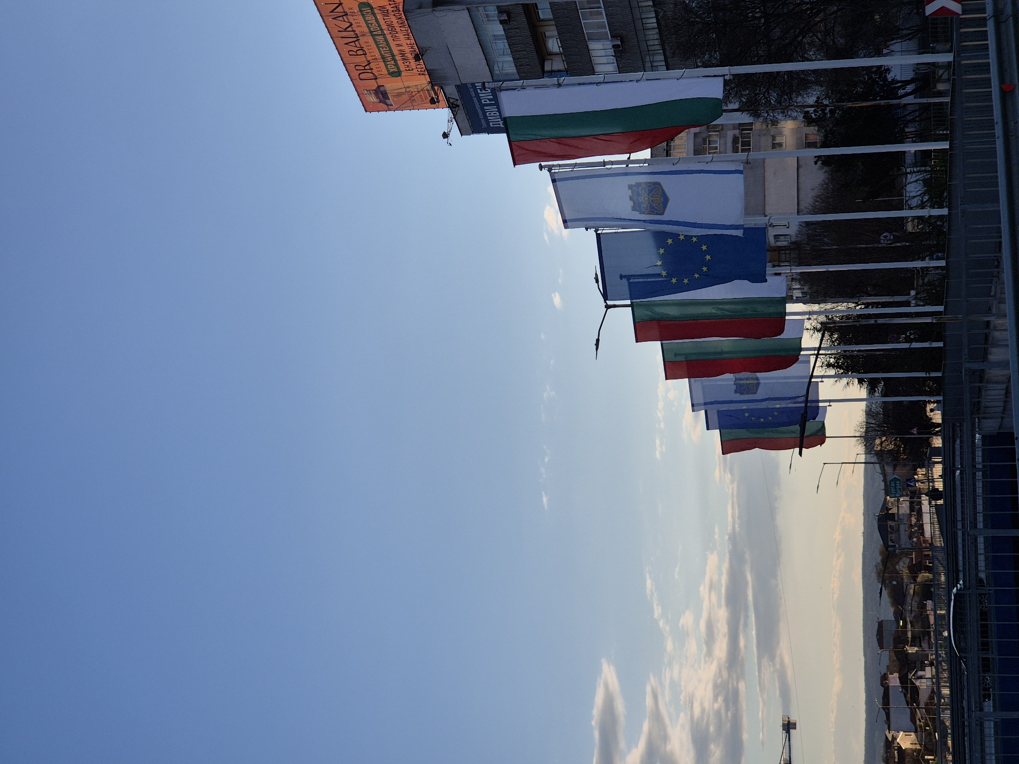 The image shows a row of flags, seemingly national and organizational, flying on flagpoles in an urban setting. The flags include the flag of Bulgaria, the flag of the European Union, and possibly the flags of local regions or cities. The photograph is taken in Varna, Bulgaria, as indicated by a sign with Bulgarian text. A building with a billboard advertising "Dr. Balkan" is visible in the upper left corner. The background shows a cityscape with buildings, trees, and a sky with scattered clouds. The camera appears to be tilted, as the sky fills a large portion of the frame. There is a metal barrier or fence on the right side of the image. The photograph was taken during daytime with good lighting. A construction crane is seen in the far background.