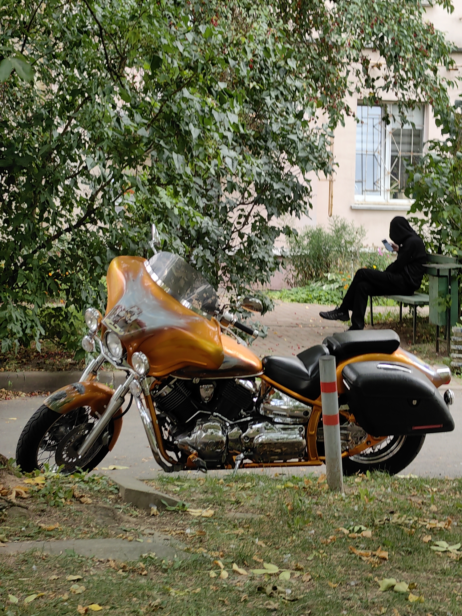 A custom orange and black motorcycle is parked on a paved surface next to a tree-lined walkway. In the background, a person wearing a black hooded sweatshirt sits on a green park bench, looking at their phone. The scene appears to be in a residential area, possibly a courtyard, given the presence of trees, a building with a window, and fallen leaves. The building is light pink and has a barred window. The motorcycle has a large windscreen and saddlebags. A concrete curb is visible in the foreground, with scattered dry leaves on the ground. The overall impression is one of an urban setting during autumn.