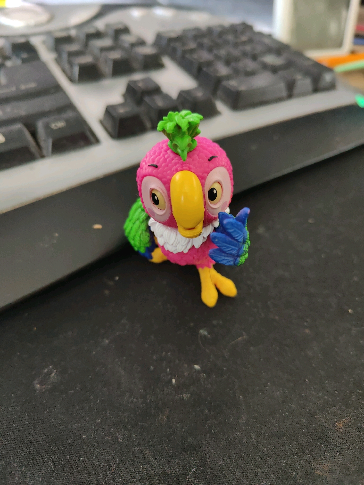 A pink toy parrot with green and blue accents sits on a black textured surface. The parrot has a large yellow beak, an eye with a black pupil, and a green tuft of feathers on its head. Its right wing is raised, and its blue hand is making a thumbs-up gesture. In the blurred background, a portion of a computer keyboard is visible. The overall scene appears to be an indoor setting, likely a desk. There is no indication of the city of Homyel, Belarus.