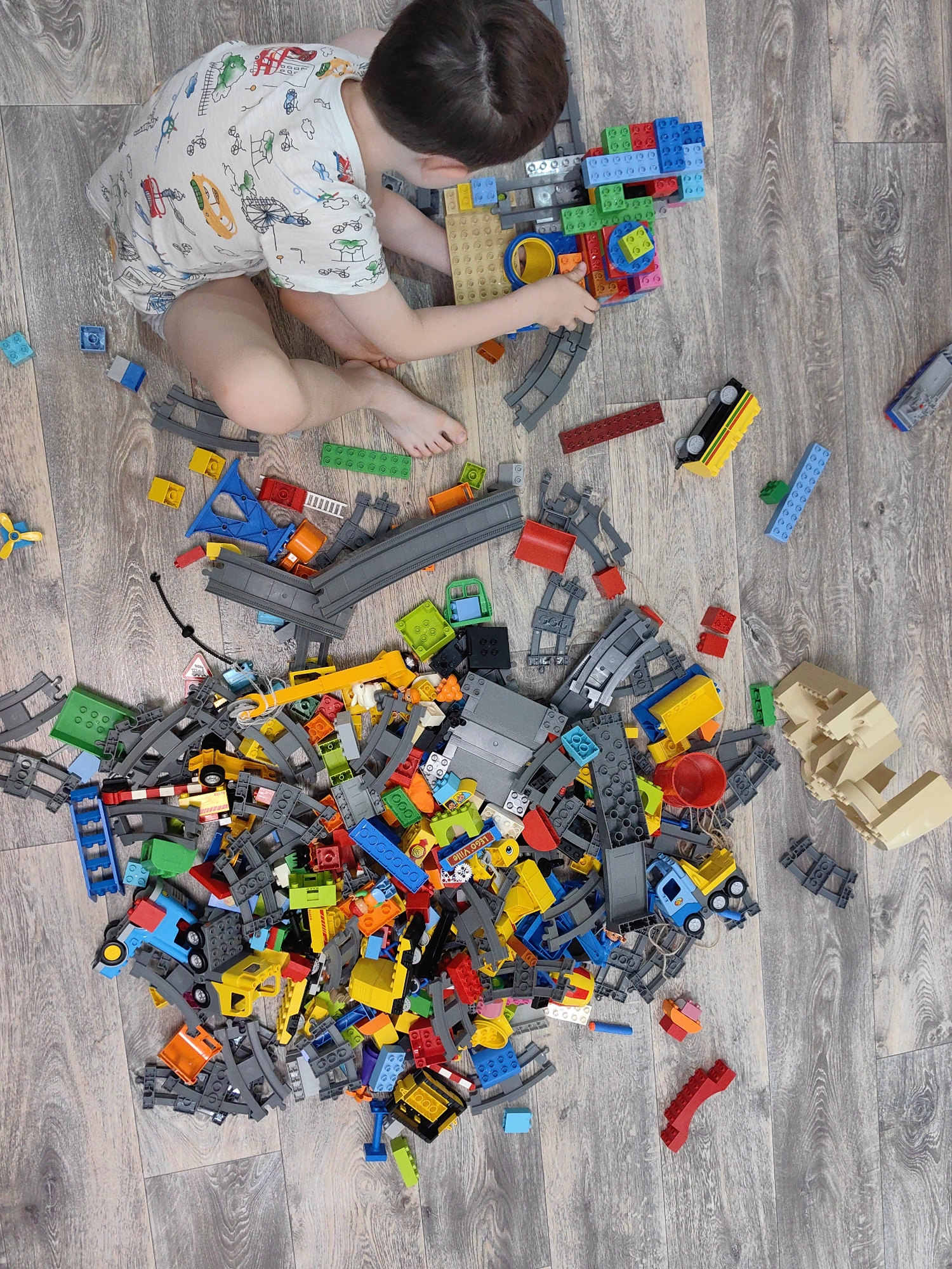 In an overhead shot, a young boy with dark hair kneels on a wooden floor, playing with a large pile of colorful building blocks and train track pieces. He wears a white t-shirt adorned with a pattern of cartoon vehicles. The blocks are scattered across the floor, with a focus on red, blue, yellow, green, and grey pieces, many of which are interlocking plastic bricks. Train track segments, some curved and some straight, are interwoven with the blocks, suggesting a construction project. A few toy vehicles are visible within the pile, including a yellow train car, a blue truck, and a small yellow propeller toy. The overall scene is one of focused play and creative construction. The image does not provide any contextual information regarding the location in Sochi, Russia.