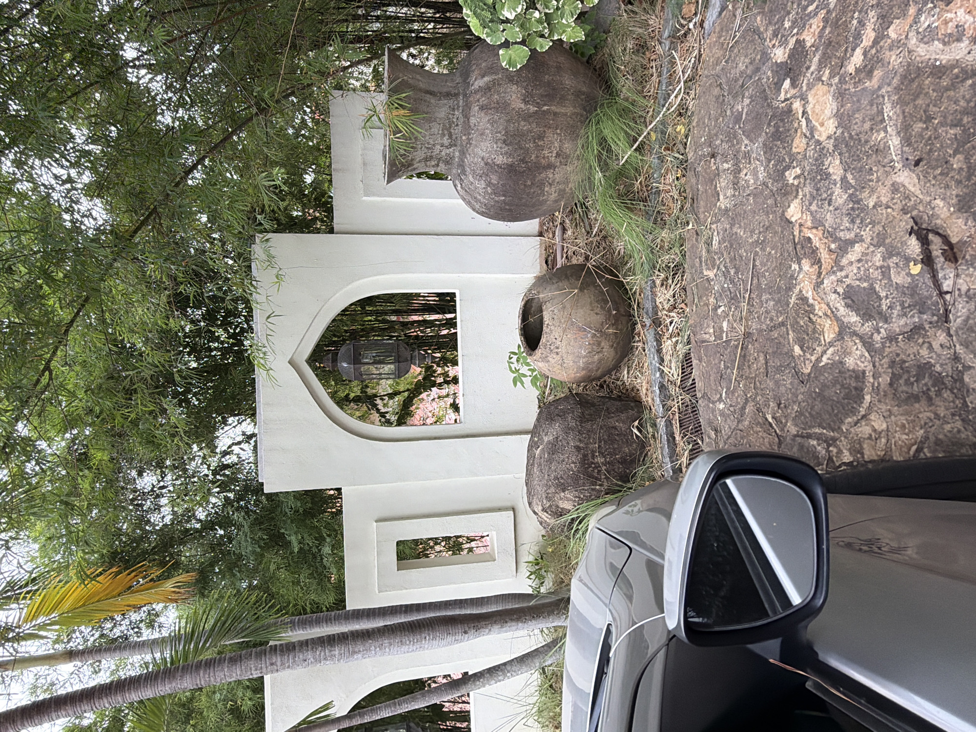 In Ko Samui District, Thailand, a grey car is parked in front of a low white wall with arched openings. Beyond the wall, bamboo and palm trees create a dense green canopy. Two large, weathered clay pots and a rough stone sit in the dry grass and straw. A large, dark-colored vase stands to the right of the scene, with lush green foliage growing nearby. The overall scene suggests a tranquil, outdoor setting with a blend of natural and decorative elements.