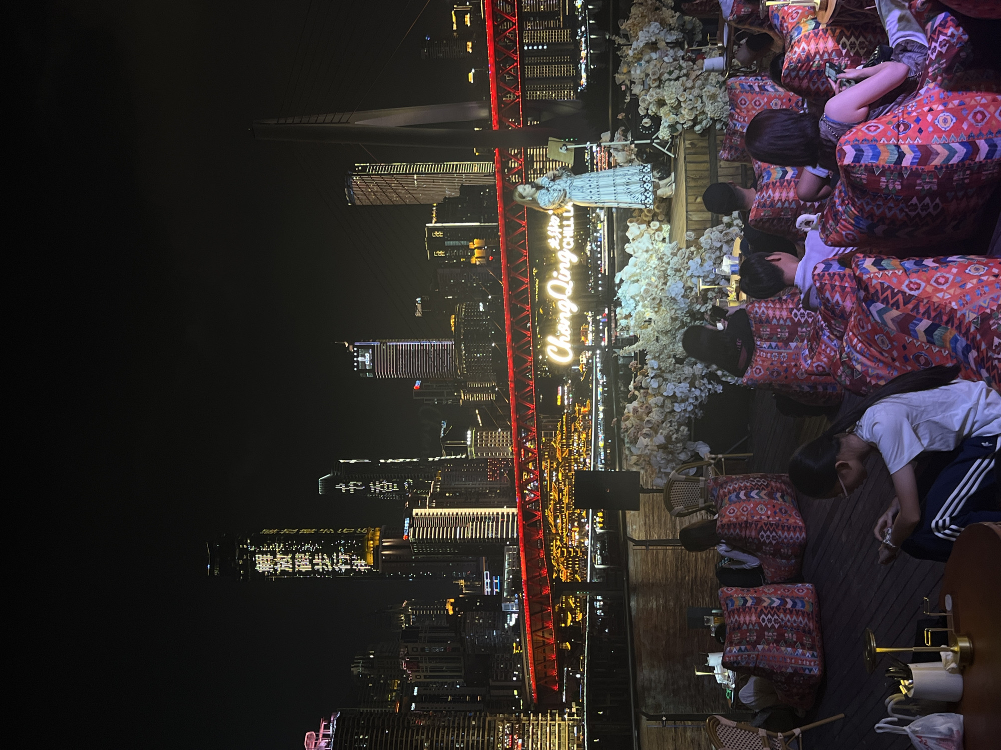 This nighttime image captures a lively scene on a wooden deck overlooking the river and city skyline of Jiangbeicheng Subdistrict, China. A woman in a patterned dress stands on a stage, singing into a microphone, illuminated by spotlights and surrounded by floral arrangements. Behind her, the cityscape is ablaze with lights, featuring towering skyscrapers and a prominent red illuminated bridge. The foreground shows an audience seated on large, colorful beanbag chairs, many of them engrossed in their phones, indicating a relaxed and casual atmosphere. The river, reflecting the city lights, flows between the deck and the distant urban landscape.