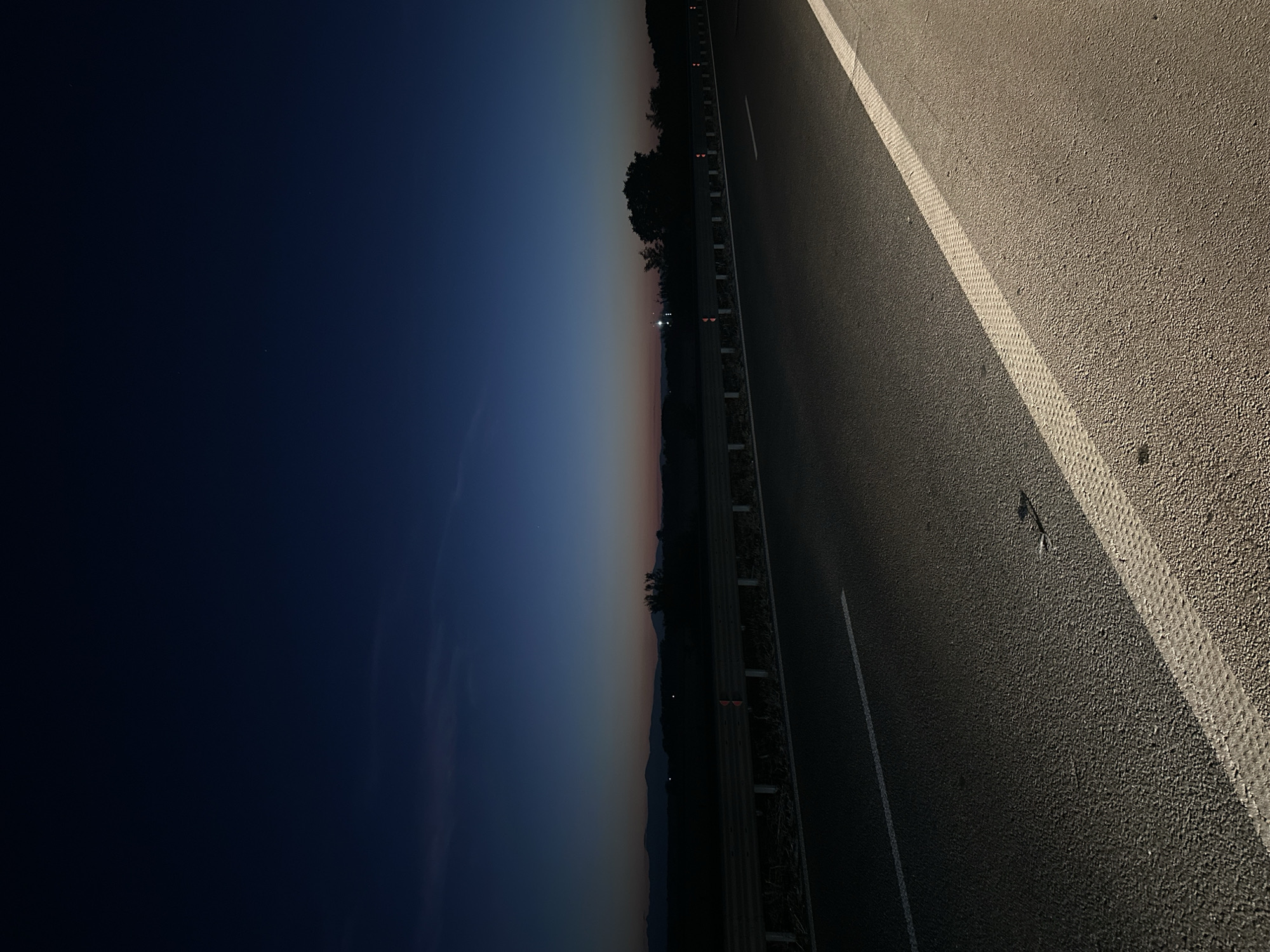 This image depicts a highway at dusk, with the sky transitioning from deep blue to lighter hues near the horizon. In the distance, a silhouetted landscape with mountains and trees is visible. The highway itself is paved, with a white line marking the lane. A guardrail runs along the side of the road, separating it from the adjacent terrain. There are no people or vehicles present in the scene. The overall atmosphere is one of tranquility and solitude.