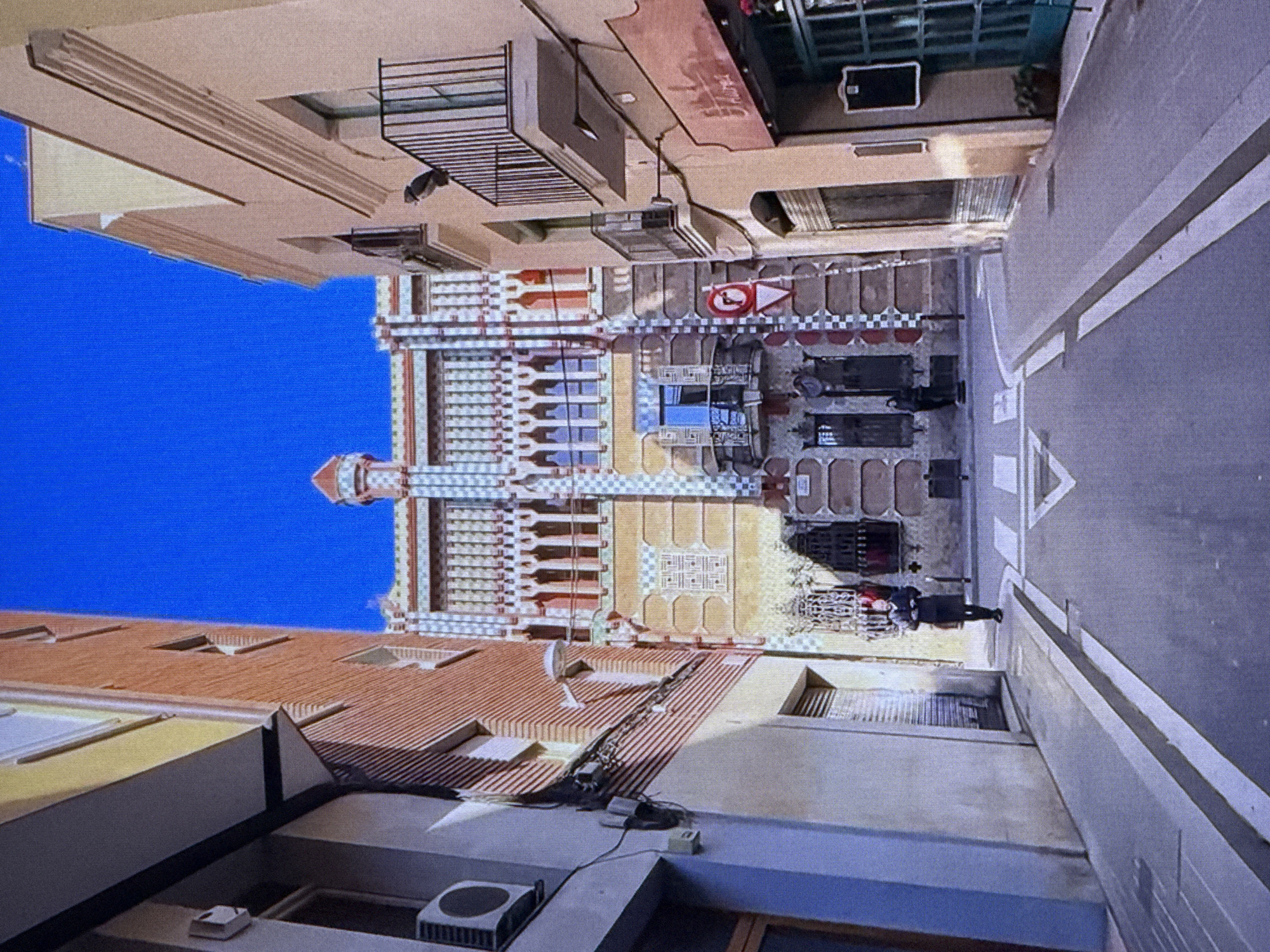 This image depicts an exterior view of a street scene with architectural elements. The primary subject is a building with a highly decorative facade, featuring intricate patterns, colorful detailing in white, yellow, red, and blue, and what appears to be a tower-like structure. The building's design suggests an artistic or historical architectural style, possibly Moorish or Art Nouveau influenced.
In the foreground and to the sides, other buildings are visible. One building on the left has a light-colored facade with layered horizontal details. Another building on the right, closer to the viewer, has a textured, light grey facade and appears to be a modern structure. The street itself is paved and features a white-lined pedestrian crossing.
There are a few visible objects, including what looks like an outdoor air conditioning unit on a rooftop, some electrical wiring, and outdoor seating or tables set up on the sidewalk in front of the ornate building. A traffic sign with a red circle and a white arrow pointing right is also visible.
Two individuals are present in the scene. One person is standing near the ornate building, partially obscured by shadow, and another person is walking away from the camera, dressed in dark clothing.
The scene appears to be set during the daytime, given the shadows cast and the bright blue sky. The weather seems clear and sunny.
There is no visible text in the image that can be deciphered. The location context provided is Dubai, United Arab Emirates.