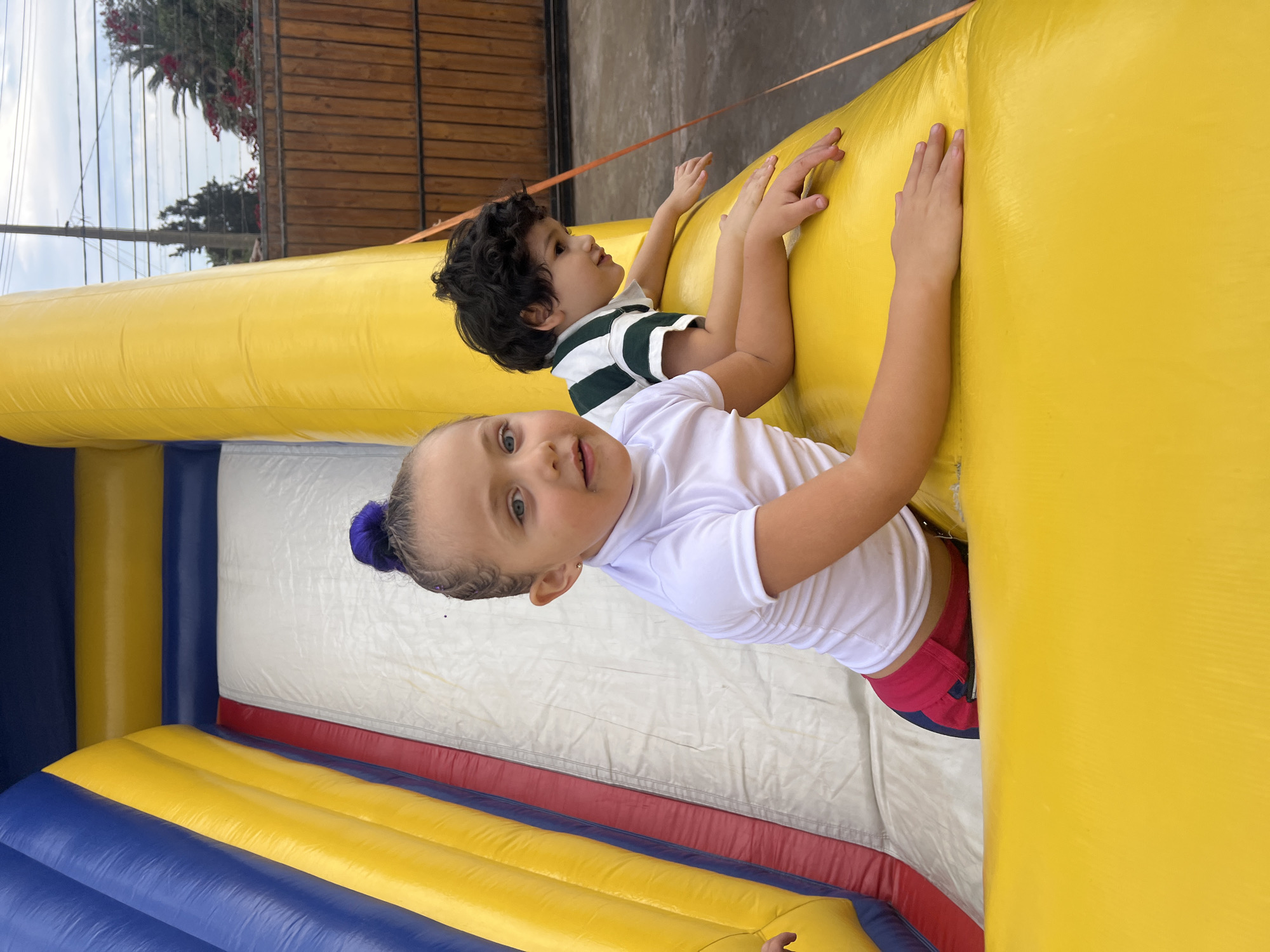 Two children are inside a brightly colored inflatable bounce house, likely in Arica, Chile. The bounce house is primarily yellow, with blue and red accents on the bottom. A girl with blonde hair pulled back in a ponytail with a blue scrunchie is in the foreground, wearing a white t-shirt and red shorts. A boy with dark curly hair and a green and white striped shirt is positioned behind her, slightly obscured. Both children are reaching out, grasping the yellow walls of the bounce house. The background includes an exterior wall and a glimpse of foliage.