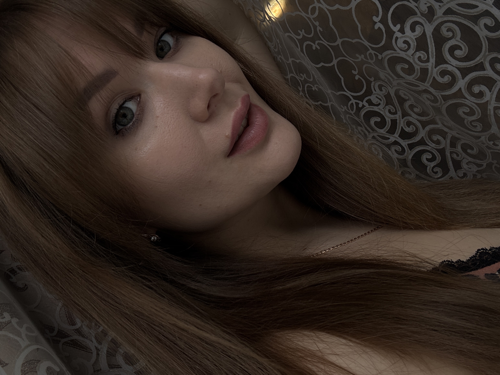 A close-up selfie of a young woman with long, auburn hair and green eyes. She is wearing a necklace and a pink and black lace top. She is lying down, with her head resting on a patterned fabric. The background is a curtain. The image appears to have been taken indoors, possibly in a bedroom. There is no indication of the location within the city of Chaikovsky, Russia.