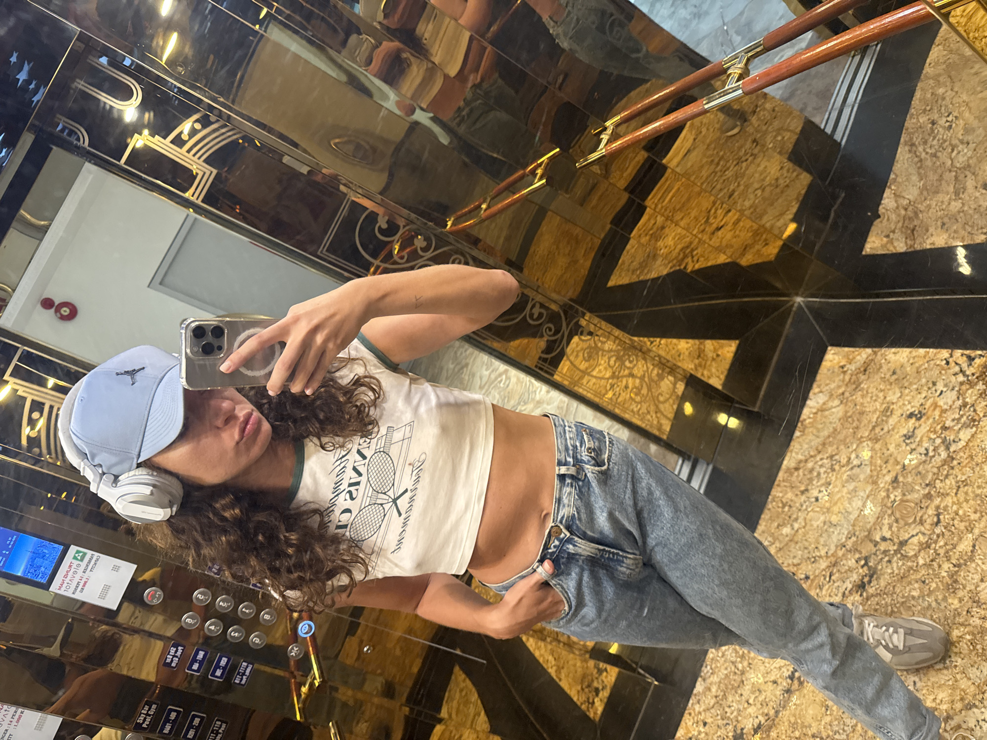 A person wearing a backwards light blue baseball cap, white wireless headphones, a white crop top with a tennis racquet graphic, light blue jeans, and gray sneakers takes a selfie in an elevator with gold walls and a black and gold floor, the elevator buttons are visible. Đà Nẵng, Vietnam is visible on one of the elevator information screens.