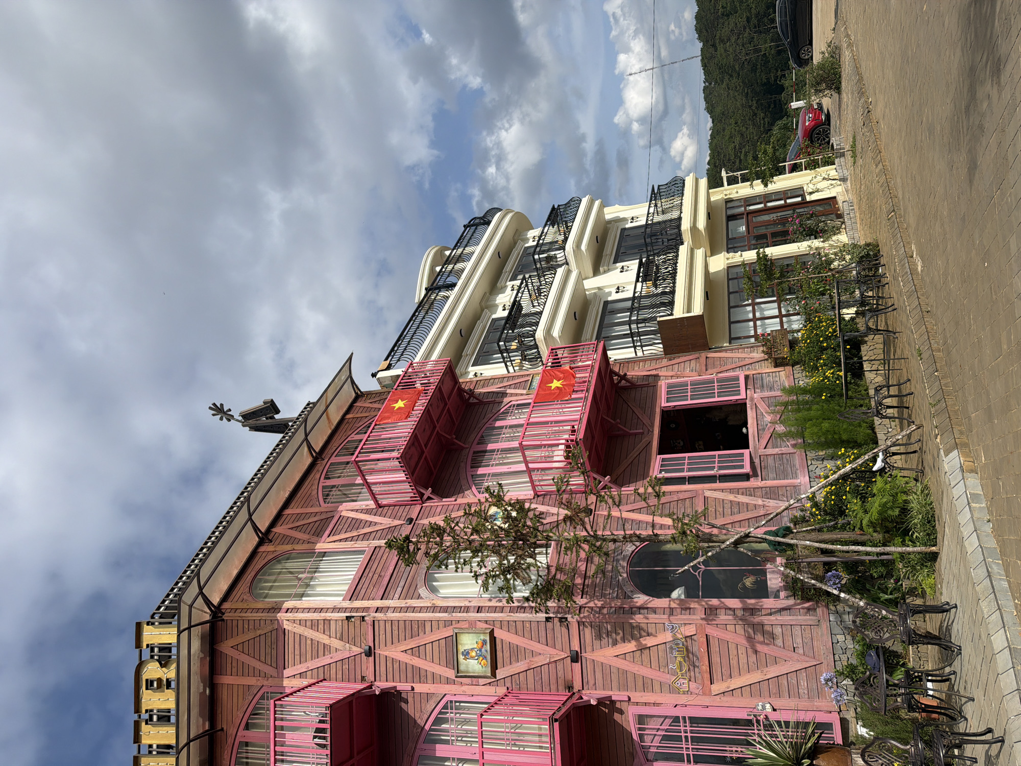 This image, taken in Măng Đen, Vietnam, captures a unique, multi-story building with a whimsical architectural style under a partly cloudy sky. The prominent structure in the foreground features a reddish-pink wooden facade composed of irregularly arranged planks and cross-braces, giving it a rustic yet playful appearance.
The building displays a variety of window shapes, including arched, round, and rectangular ones, some fitted with blinds. Several distinct pink-colored, box-like balconies protrude from the facade, two of which prominently display the national flag of Vietnam (red with a yellow star). The roof, which appears to be dark, possibly metal or tiled, is adorned with a decorative weather vane or sculpture on its peak. A partially obscured golden sign, seemingly starting with the letter "B", is visible on the left side of the roofline.
Adjacent to this distinctive building, to its right, stands a more conventionally designed multi-story structure, painted in a light cream or yellow color, featuring modern balconies with black wrought-iron railings.
The ground level in front of the pink building includes a lush garden area with various plants, shrubs, and flowering bushes, including yellow and purple blooms. Several dark metal outdoor tables and chairs are arranged on a paved brick or cobblestone surface, suggesting an outdoor seating area. A small, coniferous tree grows directly in front of the pink building.
In the background, a verdant, tree-covered mountain or hill rises, indicating a natural landscape surrounding the development. A dark-colored SUV and a red car are parked further down the paved road. The sky above is partly cloudy, with patches of bright blue visible amidst grey clouds, suggesting daytime with fluctuating sunlight.