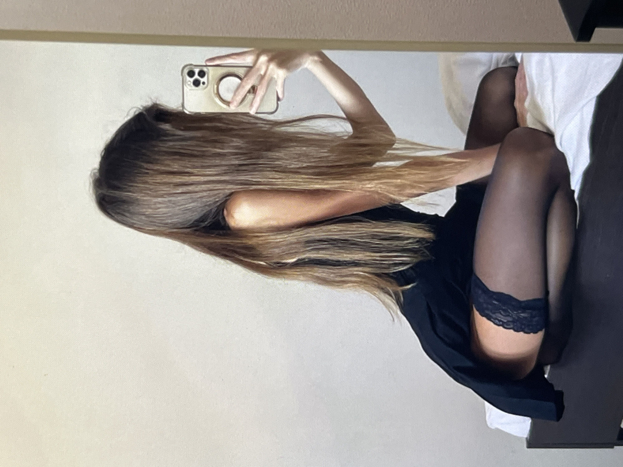 A woman with long, brown hair is taking a mirror selfie while sitting on a bed. She is wearing a black dress and black thigh-high stockings with lace trim. Her right arm is extended, holding a smartphone. The background appears to be a plain, light-colored wall. The setting is indoors, likely a bedroom.