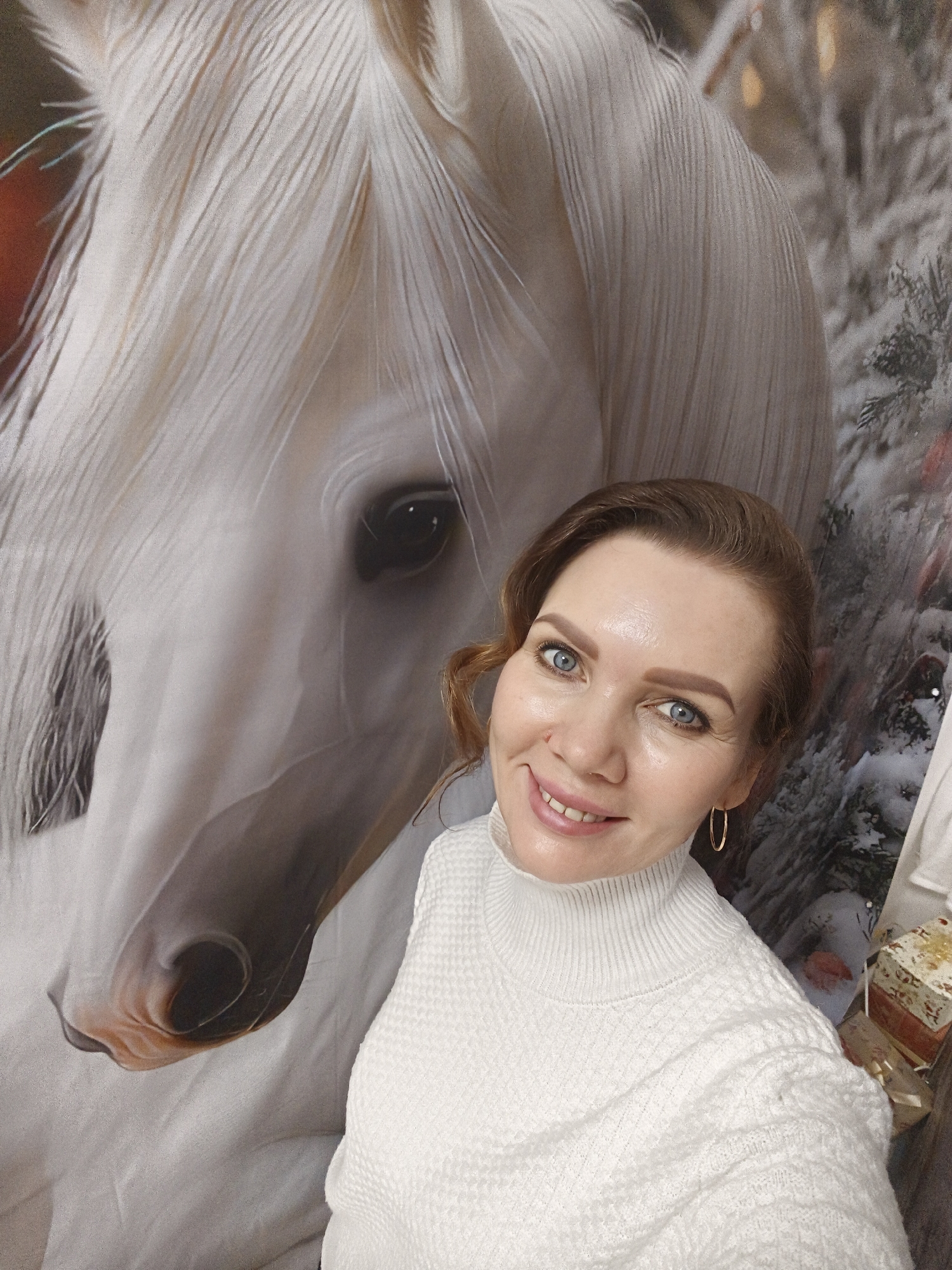 A woman poses for a selfie indoors. The background features a wall covering with an image of a white horse's head and a wintry forest scene. The woman is wearing a white turtleneck sweater and gold hoop earrings. She is smiling at the camera. The city and region are unknown.