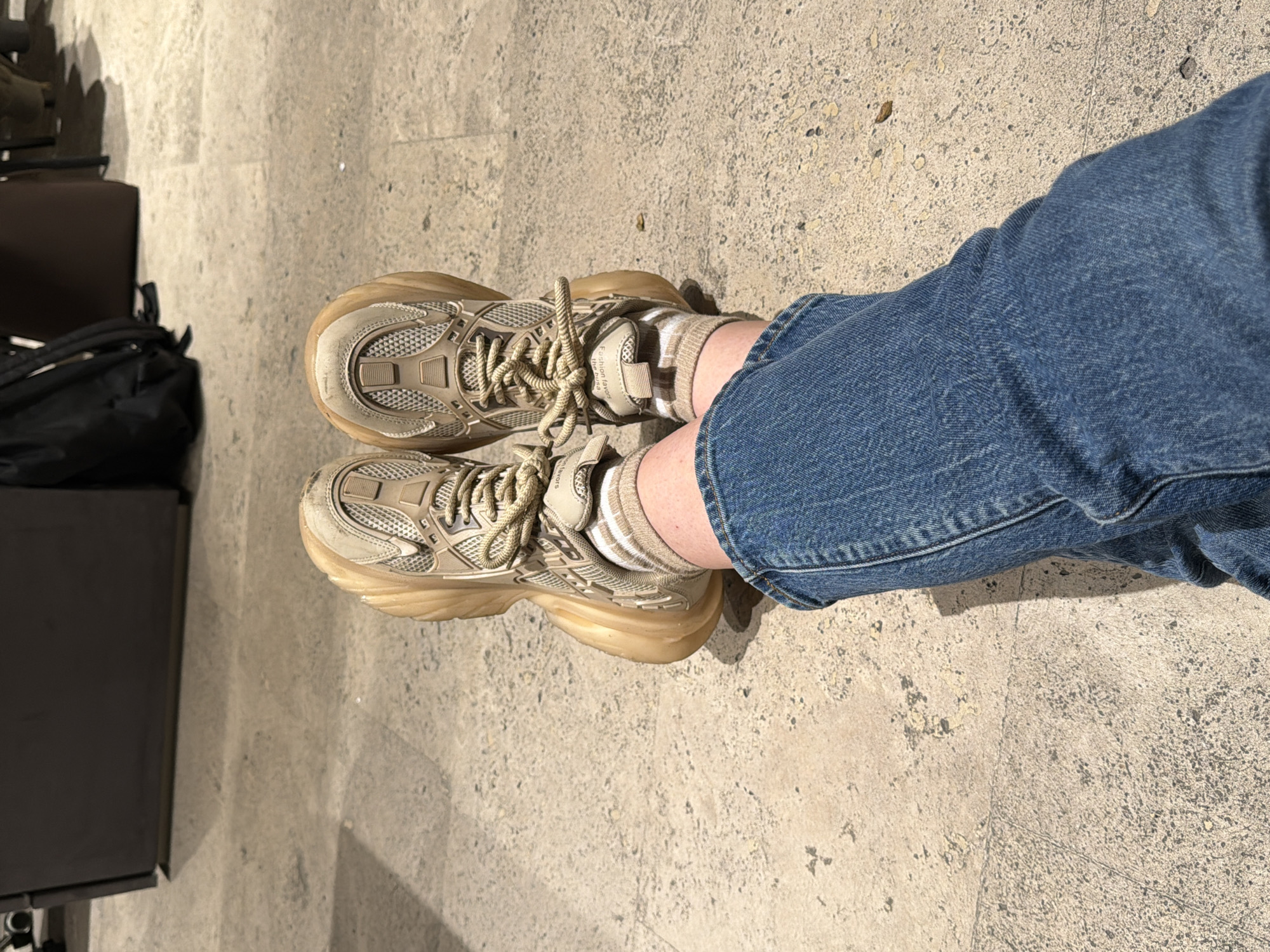 The image shows a person wearing light brown sneakers with thick translucent soles, tan socks with white stripes, and blue jeans. The person is standing on a beige concrete surface. To the left, a black backpack and a black chair leg are partially visible. The location is in Yerevan, Armenia.
