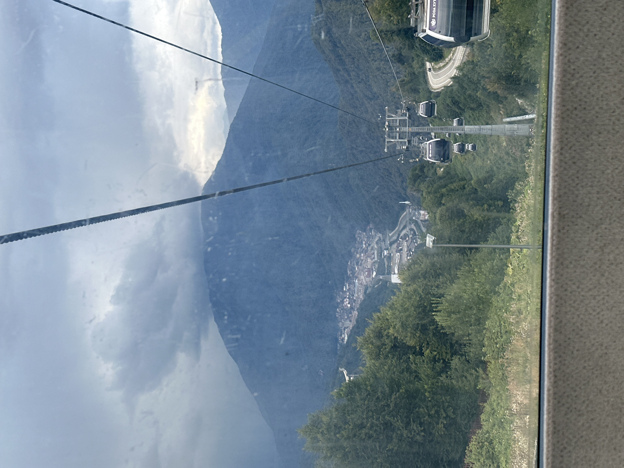 The image shows a view from a cable car moving along a ski lift in Estosadok, Russia. In the foreground, the interior of the cable car is visible, with a window looking out onto a mountain landscape. The cable car itself is suspended from a thick cable running diagonally across the frame. Below, several other cable cars can be seen ascending or descending the mountain.
The landscape consists of steep, forested mountains. A town or village is nestled in the valley below, with buildings and roads visible. A winding road with a few vehicles can be seen on the side of one of the mountains. The sky is overcast with dark clouds, suggesting either an impending storm or a generally cloudy day. The overall impression is one of a mountain resort setting. The text "РОЗА ХУТОР" is visible on one of the cable cars, which is the name of a popular ski resort in Estosadok.