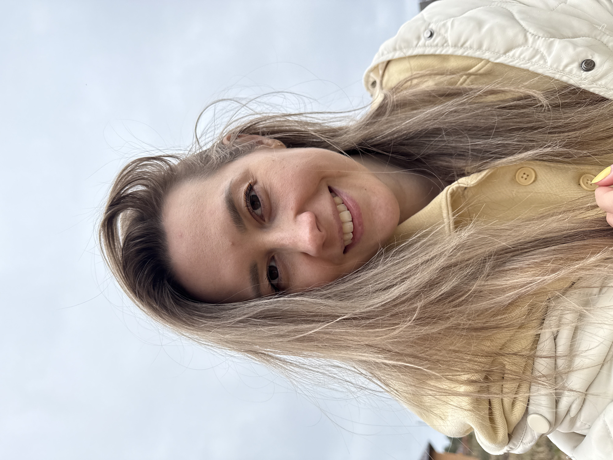 A young woman is captured in a close-up, low-angle shot, smiling broadly with her head tilted to the side. Her long, light brown hair, appearing slightly windswept, frames her face and cascades around her shoulders. She has dark eyes and a cheerful expression, revealing her teeth as she smiles. She is wearing a light yellow hooded sweatshirt, which is visible beneath a cream or off-white quilted jacket that features snap buttons. One of her fingers, sporting bright yellow nail polish, is visible near the front of her jacket. The background is a bright, plain, light blue or white sky, suggesting an outdoor setting during daytime with either clear or overcast conditions in Chaikovsky, Russia. No text is visible in the image. The overall mood conveyed is happy and casual.