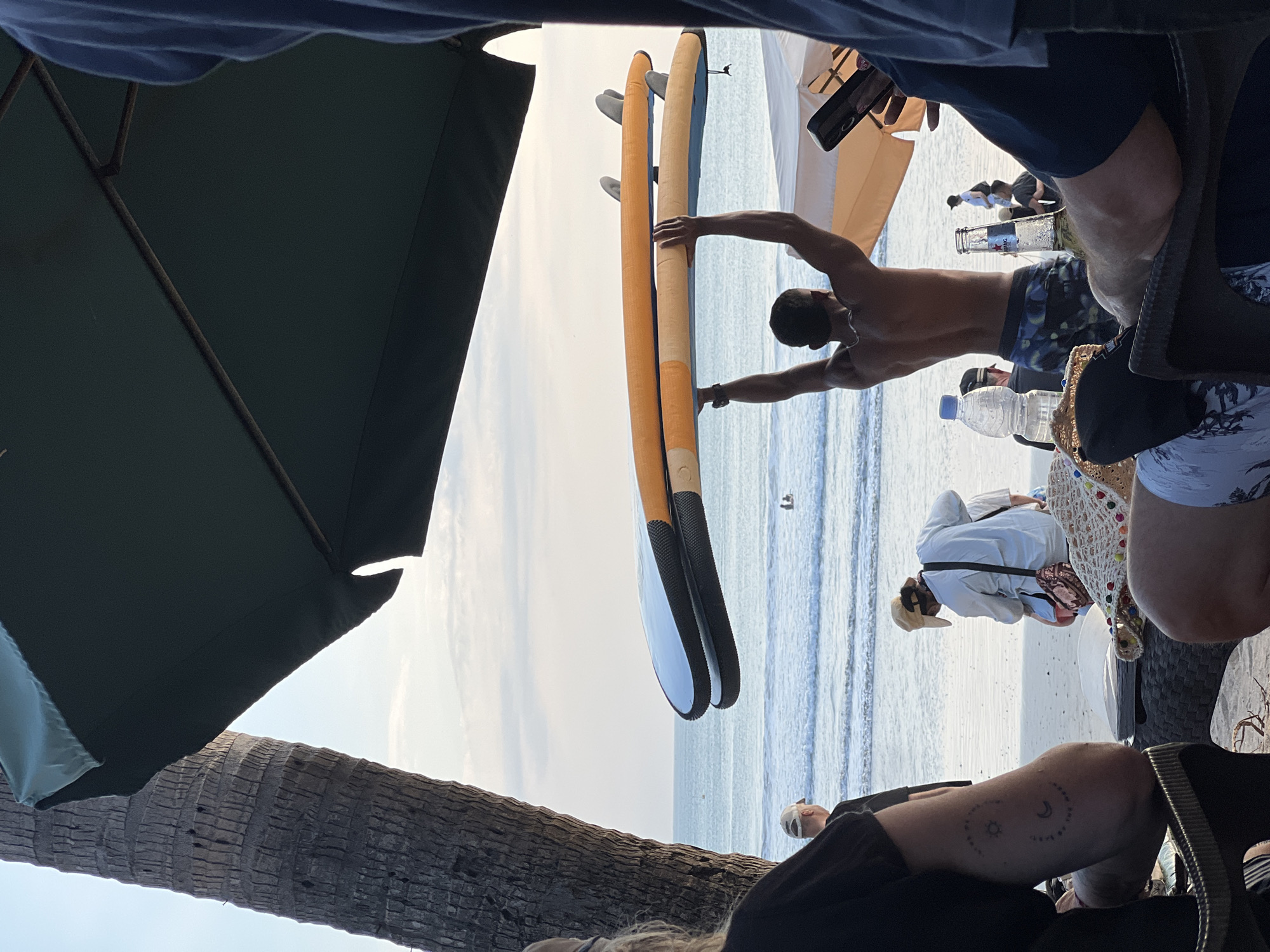 This image captures a scene on Kuta Beach, Indonesia, during what appears to be late afternoon or early evening. The foreground is dominated by people seated on lounge chairs, with a prominent palm tree trunk on the left. A man in patterned swim trunks is seen from the back, holding two surfboards above his head. His muscular physique is visible as he faces the ocean.
In the middle ground, the ocean stretches out with gentle waves. A few people are visible in the water. The beach itself is sandy, with several umbrellas and beachgoers scattered around. One person, wearing a cap, is bent over, possibly tending to something on the sand.
The background consists of the calm ocean and a hazy sky, suggesting the sun is low on the horizon or obscured by clouds. The overall atmosphere is relaxed and typical of a beachside resort area. The image appears to be taken from a slightly elevated position, looking towards the sea.