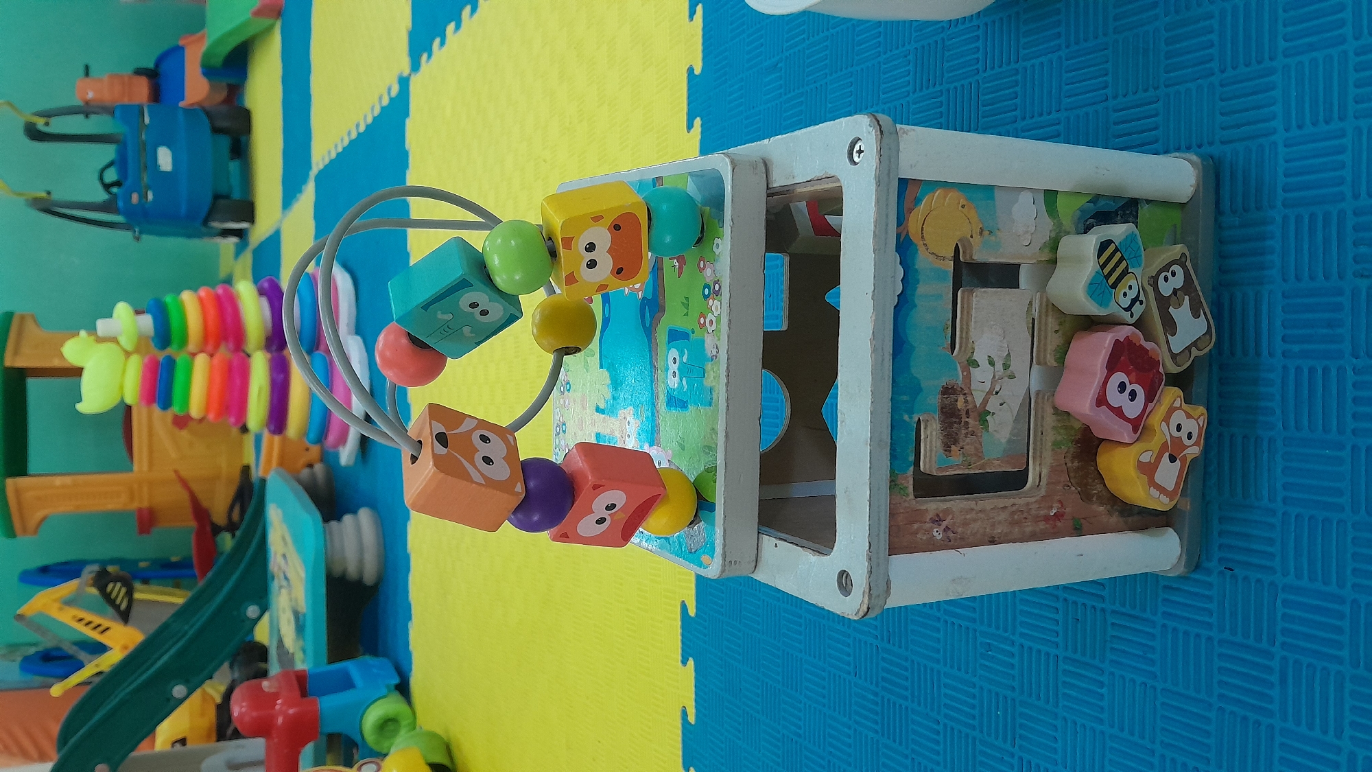 The image shows a close-up of a colorful wooden activity cube toy, possibly located in a playroom or daycare setting in Kasiman, Indonesia. The cube has various compartments and attached toys, including wooden blocks with animal faces (a fox, an owl, a bee, and a bear) and a bead maze on top with wooden cubes depicting cartoon animal faces and colored beads. The activity cube is placed on a blue interlocking foam mat, with a section of yellow foam mat visible behind it. In the background, other children's toys are visible, including a yellow and red plastic slide, a blue ride-on toy, and colorful stacking rings. The overall impression is that of a children's play area. There are no people visible in the image.