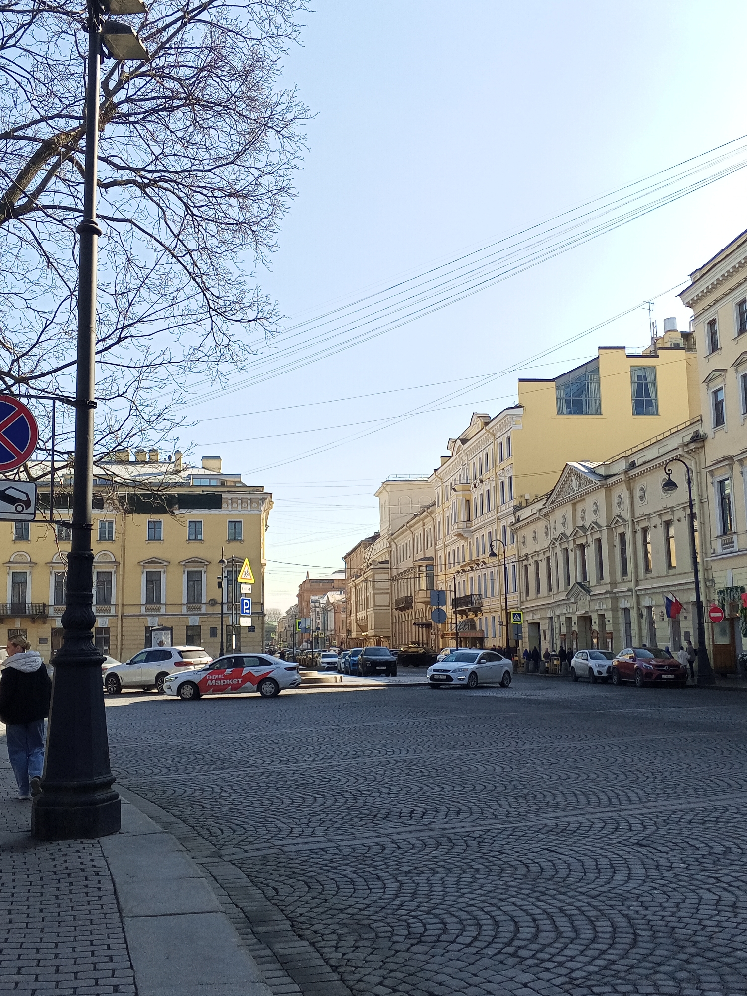 The image depicts a street scene in Saint Petersburg, Russia, characterized by cobblestone pavement and yellow buildings with ornate facades lining the street. Several vehicles are present in the intersection, including cars of varying colors and a Yandex Market branded vehicle. Street signs and a lamp post are visible on the left. A pedestrian is walking on the sidewalk to the left. Bare tree branches are seen against a light blue sky. Overhead, several power lines are strung across the scene. The buildings exhibit classical architectural styles with detailed window frames and moldings. Some pedestrians are visible on the sidewalks along the buildings.