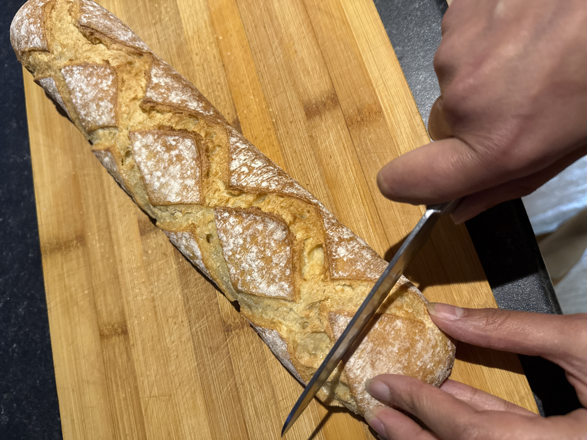 An overhead shot captures a baguette being sliced on a wooden cutting board. The baguette, dusted with flour, displays a crisscross score pattern on its golden-brown crust. A metallic knife, held by two hands, is mid-cut into the bread. The hands, with visible nails, are positioned to grip and guide the knife. The cutting board has a natural wood grain and appears to be a standard kitchen utensil. The background is dimly lit, with a dark countertop visible at the top and a lighter surface below. The scene is devoid of any specific Great Yarmouth landmarks or indicators.