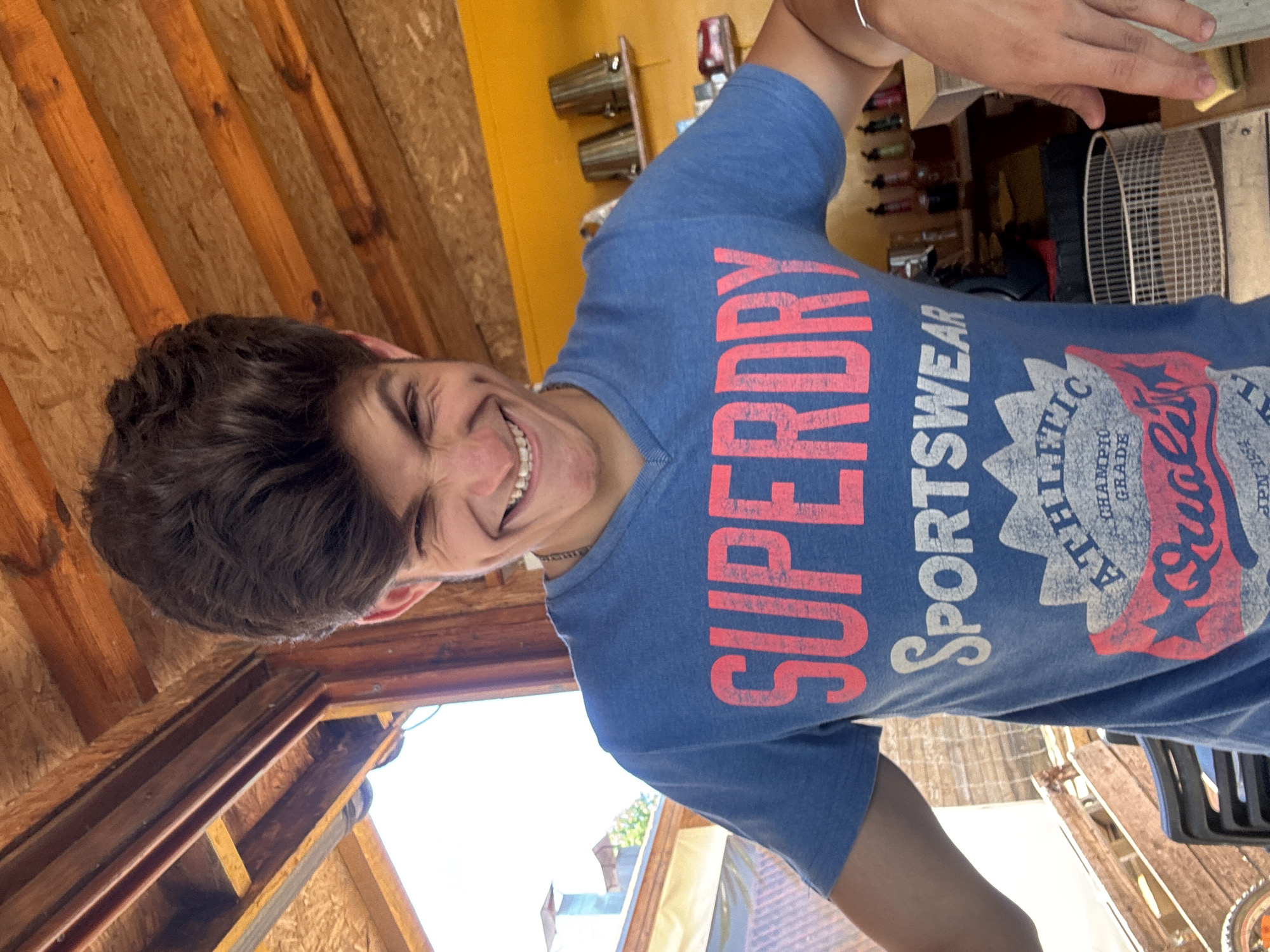 A young man with dark, voluminous hair and a wide, toothy grin is captured in a close-up shot. He is wearing a blue t-shirt with a graphic design that includes the words "SUPERDRY SPORTSWEAR ATHLETIC QUALITY GRADE". The man's left arm is bent at the elbow, with his hand resting on his hip. His right arm is partially visible, with a silver bracelet on his wrist.
The background suggests an outdoor setting with wooden beams and yellow walls. On the wall, there are shelves with various items, including what appear to be metal containers and bottles. The lighting indicates it is daytime, with bright sunlight casting shadows. The environment appears to be rustic, possibly a cafe or an outdoor seating area.
This scene does not contain any elements that would allow for a specific identification of the location within the city of Sozopol, Bulgaria.