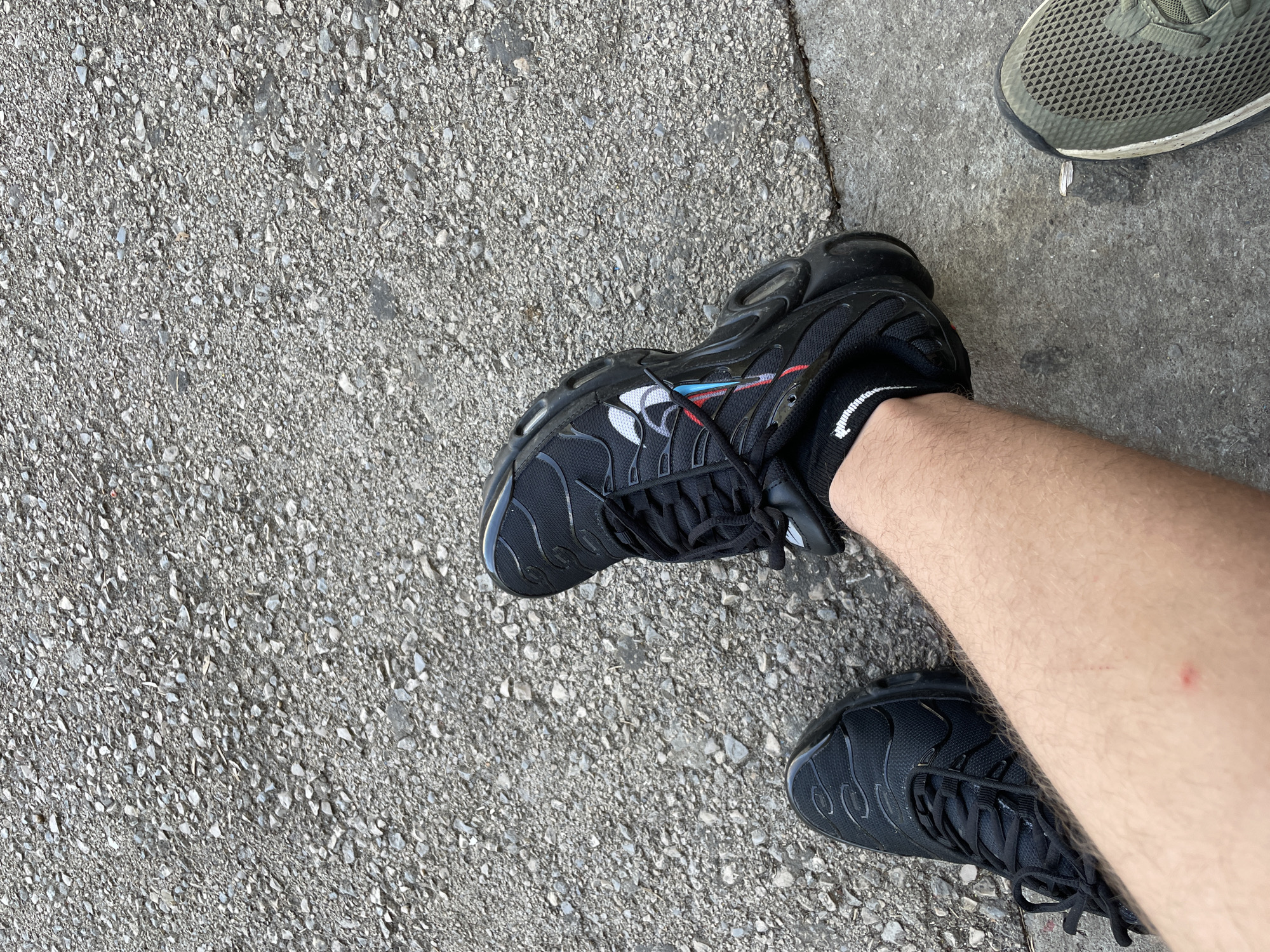 An overhead, slightly angled shot captures a person's lower legs and feet on a textured grey ground surface in Troyan, Bulgaria. The person is wearing black sneakers, identified as Nike Air Max Plus (TN) models, on both feet. The left sneaker prominently displays subtle red and light blue accents on its side, near the laces, complemented by a white Nike swoosh logo. A white sock with a black stripe is visible on the left ankle, while the right leg is bare, revealing a small red mark or bite on the calf.
The ground consists of coarse asphalt or concrete, made up of various small grey and white stones. In the upper right corner of the frame, the front portion of an olive-green mesh sneaker is partially visible, suggesting the presence of another person or an abandoned shoe nearby. The lighting indicates it is daytime, with clear weather. No specific activity beyond standing or walking is discernible. No distinct legible text is visible other than the Nike swoosh on the shoe.