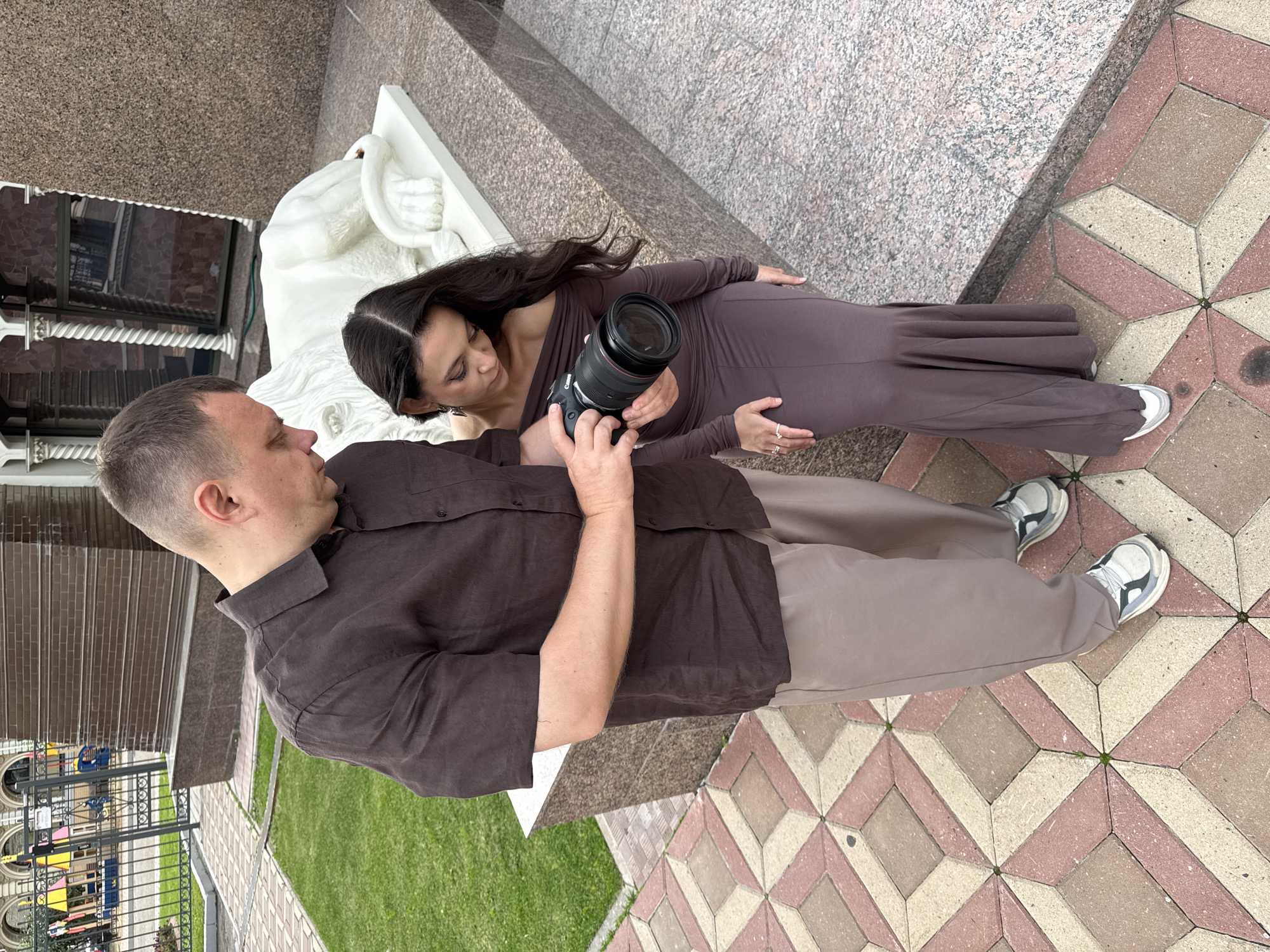 A man and a woman are standing on a patterned paved walkway outside a building. The man, wearing a brown short-sleeved shirt and light-colored pants, is holding a black camera with a large lens. He is looking down at the camera. The woman, wearing a long, off-the-shoulder brown dress, is standing next to him, also looking down at the camera. A white sculpture of a lion is visible behind them, and part of a building with a textured facade is in the background. The setting appears to be in a city, potentially Kazan, Russia, given the architectural style and paving.