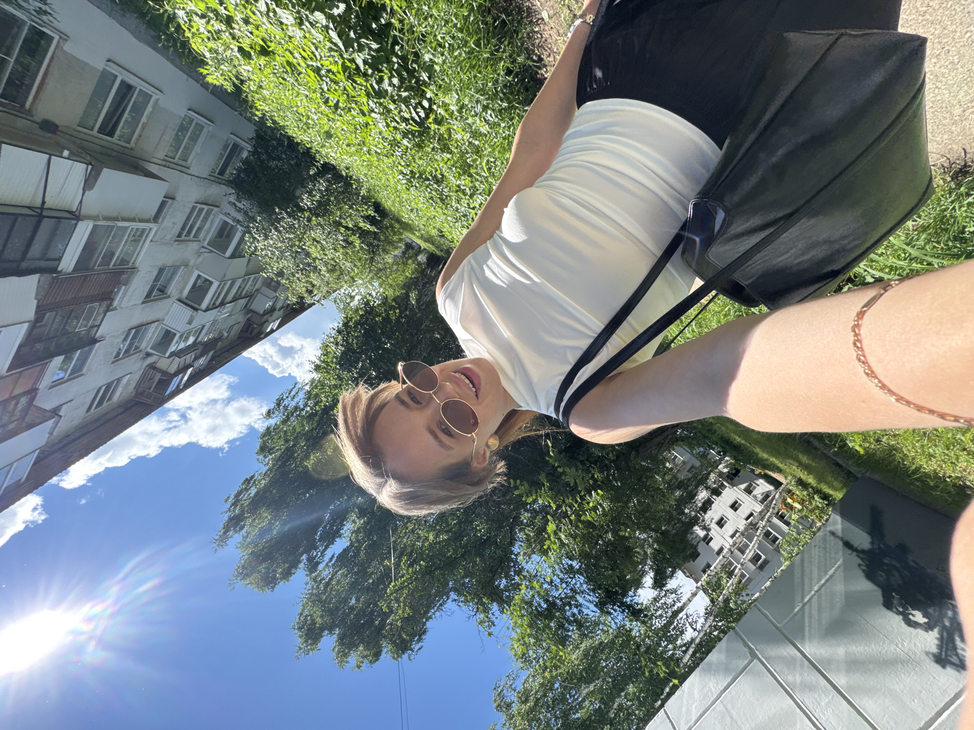 The image is an outdoor selfie taken from a low angle, looking up at a woman with round sunglasses. She wears a light-colored, short-sleeved shirt with dark trim, and black pants, and carries a black handbag. She has a gold bracelet on her wrist. The background includes a multi-story building with windows and balconies, green trees and grass, and a bright blue sky with the sun visible. The location is the city of Chaikovsky, Russia.