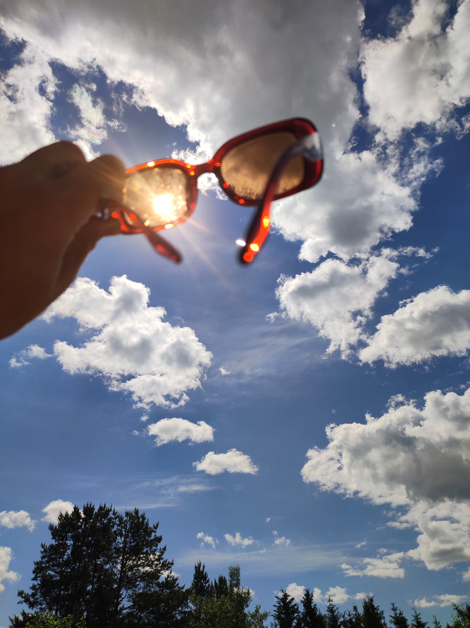 The image captures an outdoor scene from a low-angle perspective, looking upwards towards a bright sky in Фоки, Russia. In the foreground, a human hand, visible in partial silhouette and slightly out of focus, holds a pair of reddish-brown, possibly tortoise-shell, framed sunglasses. The sun is positioned directly behind the left lens of the sunglasses, creating an intense, starburst-like light flare and glare through the lens, obscuring its details and illuminating the inner frame. The sunglasses themselves are also slightly out of focus. The background consists of a vibrant blue sky populated with numerous white and grey cumulus clouds, varying in size and density, distributed across the frame. Along the bottom edge of the image, the dark, silhouetted tops of several trees, appearing to be coniferous, are visible against the bright sky, indicating foliage at the horizon. The scene is brightly lit by direct sunlight.