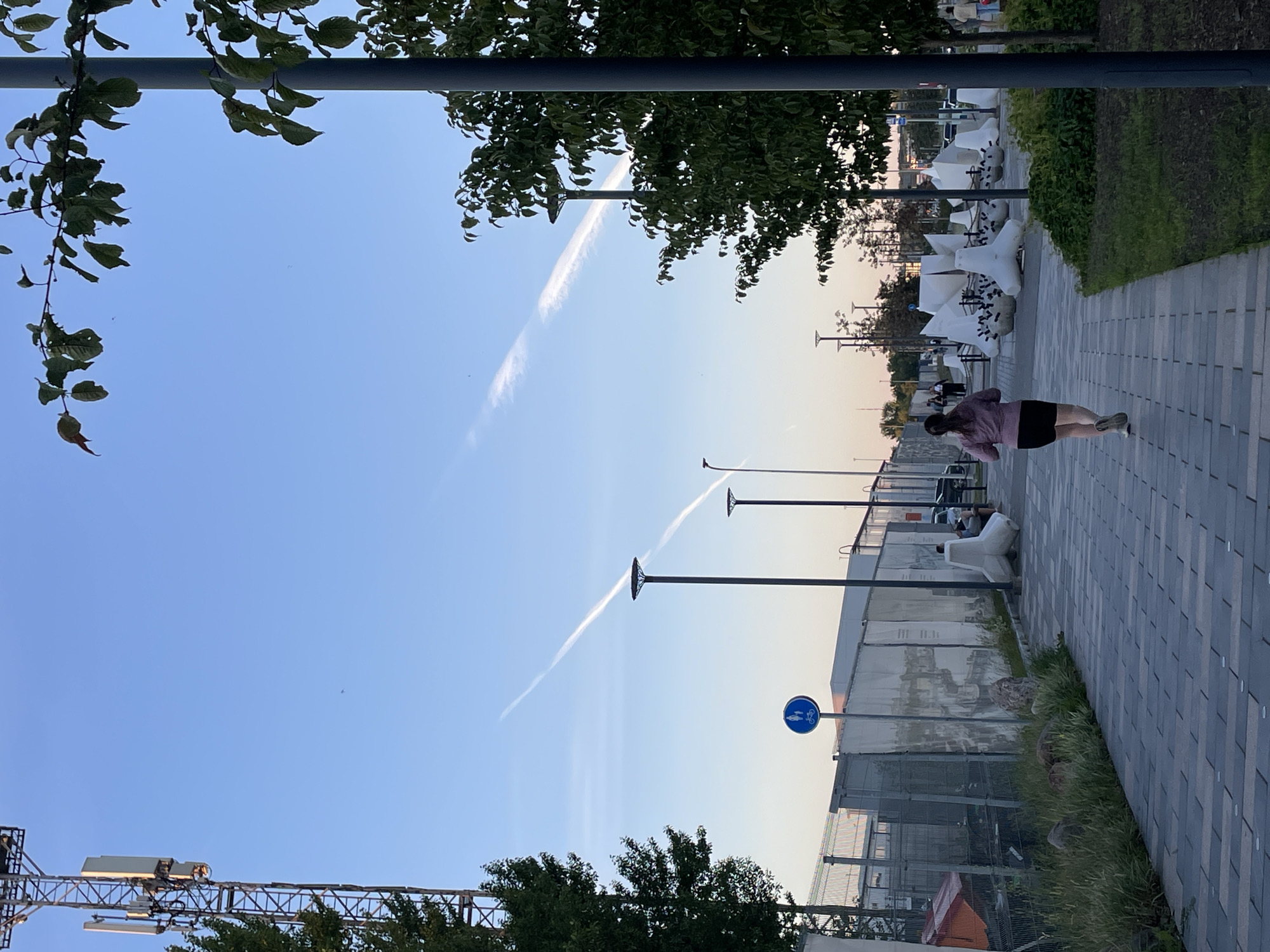 The image depicts a wide paved walkway in a city, likely an urban park or promenade, with a person jogging away from the camera. The setting appears to be Tallinn, Estonia, with a tall cell tower visible on the left. Two distinct contrails stretch across the clear blue sky, suggesting aircraft activity.
The jogger, a woman wearing a pink hoodie and black shorts, is positioned in the center of the frame on the walkway. The walkway is paved with dark grey and lighter grey rectangular tiles, laid in a patterned manner. To the left of the walkway is a fenced-off area with tall grass and some rocks. A blue circular sign with icons of a person walking and a bicycle indicates pedestrian and cyclist paths. Further left, a large cell tower with multiple antenna arrays dominates the skyline.
To the right of the walkway, a series of abstract white sculptures are arranged, some resembling wave breakers or artistic installations. Beyond the sculptures, trees with green foliage line the area. A prominent dark grey lamppost stands in the foreground on the right. In the distance, more streetlights and trees are visible, along with some parked cars and buildings. The overall atmosphere is serene, with the late afternoon or early evening sun casting a soft light.