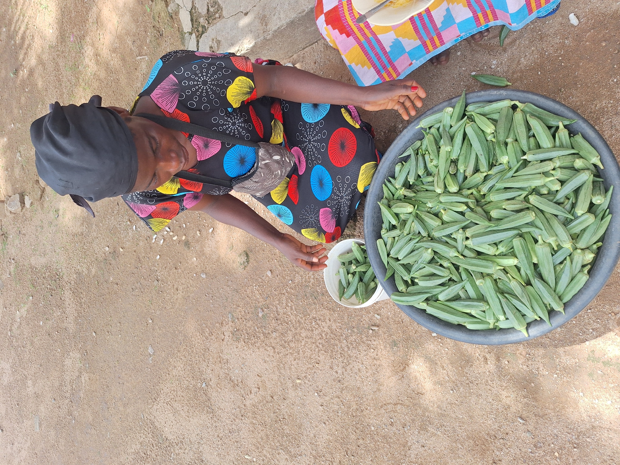 An eye-level, high-angle shot depicts a woman in Sabon Tasha, Nigeria, seated on sandy ground next to a large black bowl overflowing with okra. The woman wears a dark head covering and a colorful dress with radial patterns. A patterned cloth sits to her right. A small white bucket beside her holds a smaller quantity of okra. She is in the process of sorting or preparing the okra. The environment is open and appears to be an outdoor market or roadside setting, characterized by bare earth.