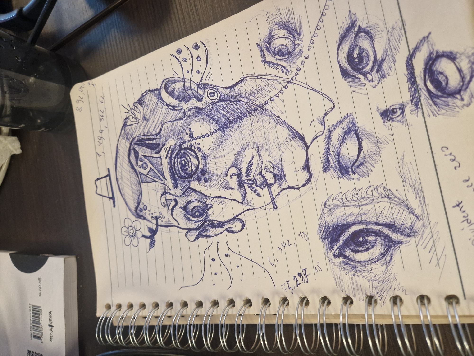 The image shows an open notebook with a spiral binding, containing pen drawings of eyes and an abstract face. Numerical notations are also visible on the page, along with the words "violent zero" near the right edge. Next to the notebook, on the left side of the image, are portions of a dark-colored water container and a white box with the words "MUSA GENA" and an ISBN number printed on it. The surface the notebook is resting on appears to be a dark-colored wooden table or desk.