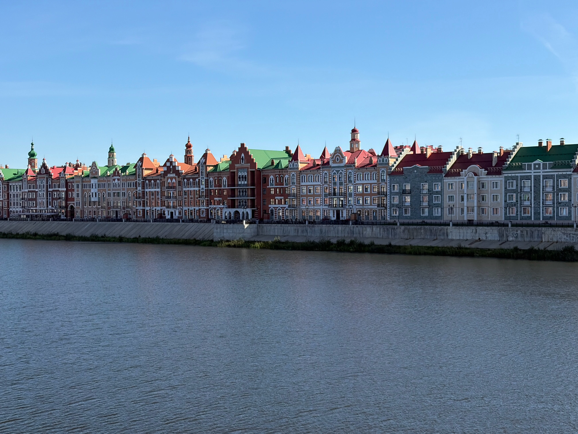 The image displays a picturesque waterfront in Yoshkar-Ola, Russia, featuring a row of colorful, European-inspired buildings with red and green roofs. These multi-story structures are characterized by their varied architectural styles, including dormer windows, decorative facades, and turrets, creating a vibrant and uniform skyline along the riverbank. A concrete embankment separates the buildings from the wide, calm river in the foreground, its surface rippled by a gentle breeze. The sky above is a clear, cloudless blue. No people are visible in the scene.