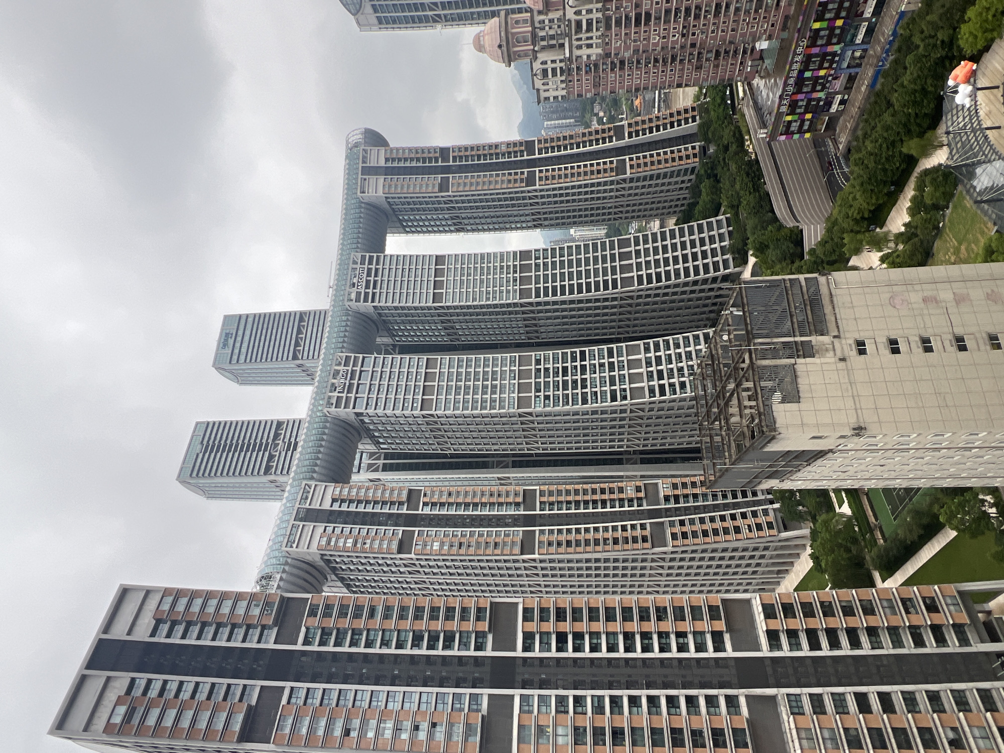 An aerial view depicts the Raffles City complex in Yuzhong District, China, a striking architectural ensemble characterized by its interconnected skyscrapers. The most prominent structure is a horizontal ribbon-like building connecting four vertical towers, its transparent canopy curving dramatically over a vast open space. The towers themselves are clad in a mixture of grey and reddish-brown panels, with numerous rectangular windows creating a geometric pattern. To the left, a cluster of residential high-rises displays a similar design, featuring dark grey and lighter colored facades interspersed with windows. In the foreground, a light-colored rectangular building with a caged roof structure occupies the lower right quadrant. Beyond the central complex, a cityscape unfolds with more buildings of varying heights and architectural styles, including a distinctly styled pink domed building. Lush green trees and landscaped areas, including a tennis court, are visible between the buildings, adding a touch of nature to the urban environment. The sky is overcast, with a dense layer of grey clouds dominating the upper portion of the image.