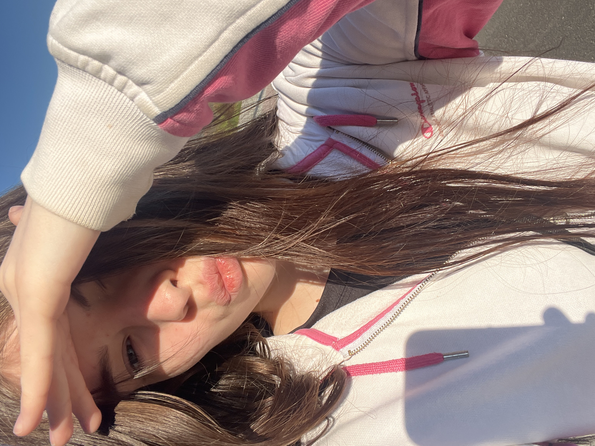 A medium close-up, rotated outdoor shot depicts a young person, likely female, with long, dark brown hair spread around them. The person is wearing a white zip-up hoodie featuring pink accents, including a pink zipper lining, pink drawstring tips, and a pink stripe on the shoulder/sleeve. The hoodie also has a visible "Champion" logo in pink, with "ATHLETIC APPAREL" printed beneath it.
The subject is looking towards the camera with a neutral expression, their lips slightly parted. One hand is partially covering their forehead and hair, adding to a casual, possibly sun-shielding pose. Strong sunlight is evident, creating clear shadows across the person's face and clothing, particularly on the hoodie and under the hand. A clear blue sky is visible in the background, suggesting a sunny day. The setting appears to be outdoors, possibly on a paved surface. The image is taken in Saint Petersburg, Russia.