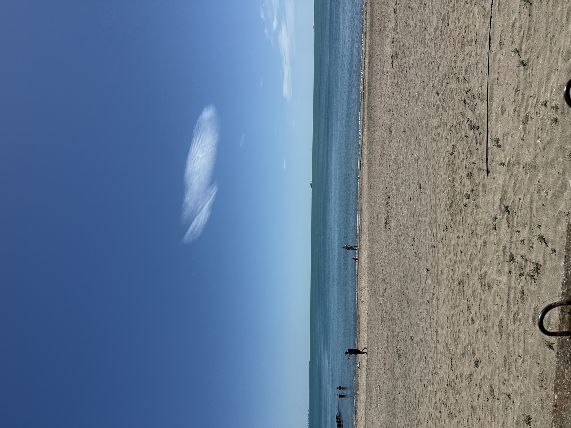This image depicts a beach scene in Burgas, Bulgaria. The sky is clear blue with a singular wisp of cloud. The sea stretches along the horizon in a muted blue. The sandy beach is marked with footprints and sparse vegetation. Several small groups of people are walking along the shoreline and wading into the water. A black metal ring can be seen affixed to the right edge of the scene.