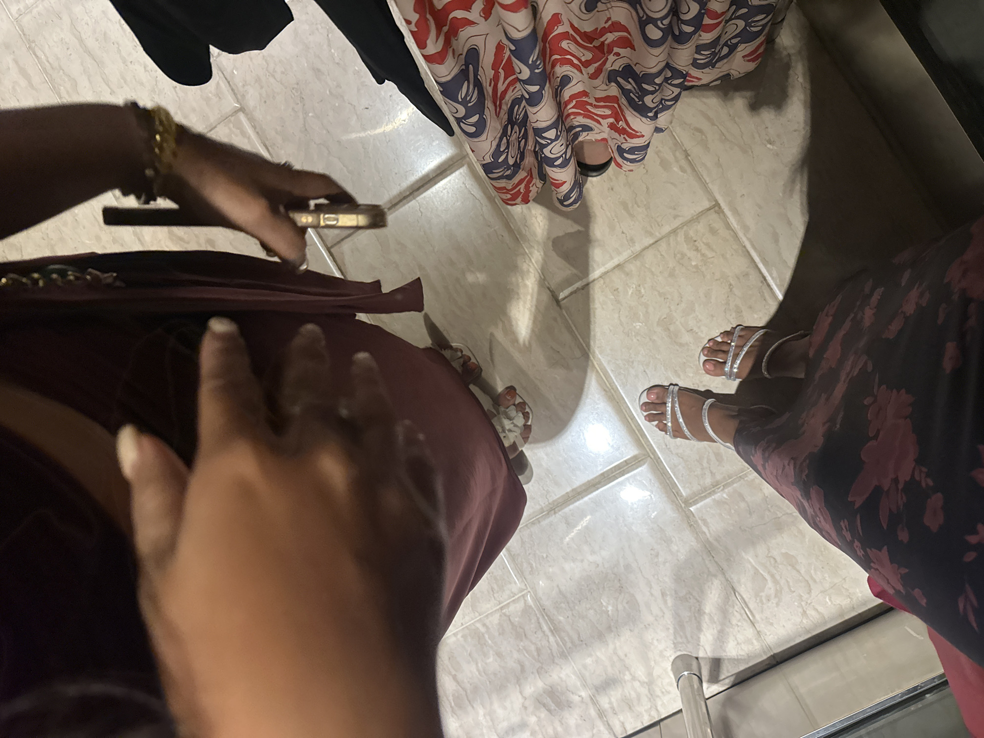 This high-angle image captures an indoor scene in Kuta Selatan, Indonesia, focusing on the lower halves of several individuals on a light-colored, possibly beige or grey, tiled floor. The floor tiles are large and have distinct grout lines, illuminated by artificial light that casts subtle shadows.
In the foreground, a person is wearing a dark maroon or deep red dress or skirt. Their left hand, adorned with multiple gold bracelets, is visible holding a gold-colored smartphone. Their right hand is closer to the camera, partially out of focus.
To the right, another individual is seen wearing a long dress or skirt featuring a dark background with a pattern of red and pink flowers. This person is wearing elegant silver strappy sandals. A shiny, metallic railing or bar is partially visible on the floor near their feet in the lower right corner.
Further into the background, slightly to the left, the lower part of a third person's dress is visible. This dress has a striking pattern of red and blue wavy or abstract motifs on a cream or light-colored fabric. This person is wearing black, closed-toe shoes. Hints of a fourth person's feet, wearing white sandals with floral details, can be discerned from the combined perspective of the crops.
The image suggests a casual gathering or an outing, possibly in a shopping center or another public building, given the varied clothing styles and the indoor setting. There is no visible text in the image.